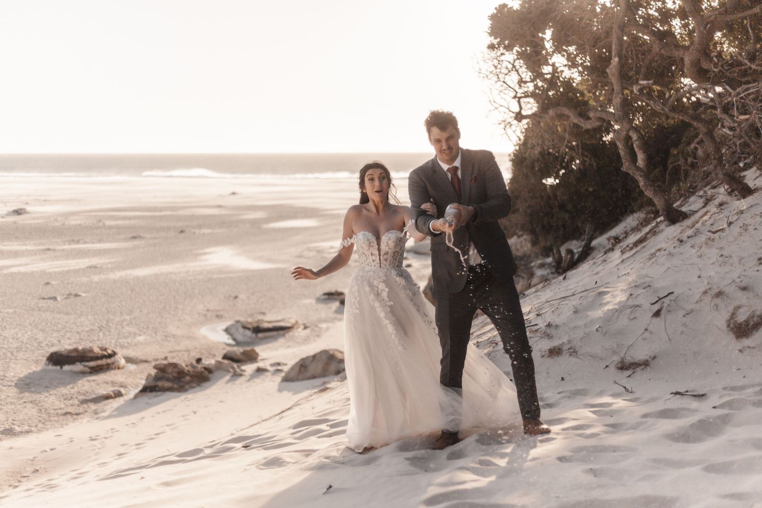 Bride and groom celebrating on sandy beach dunes, popping champagne in golden sunlight near coastal trees.