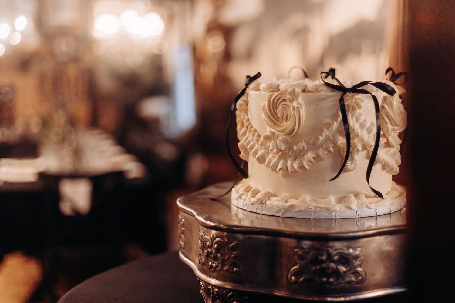 Elegant white wedding cake with ruffles and black bow ribbon on ornate silver stand in warmly lit reception venue.