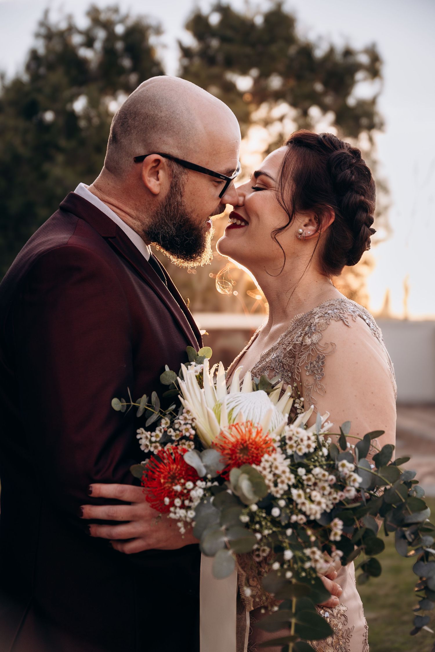 Bride and groom sharing a romantic moment, holding a bouquet of white and red flowers at golden hour.