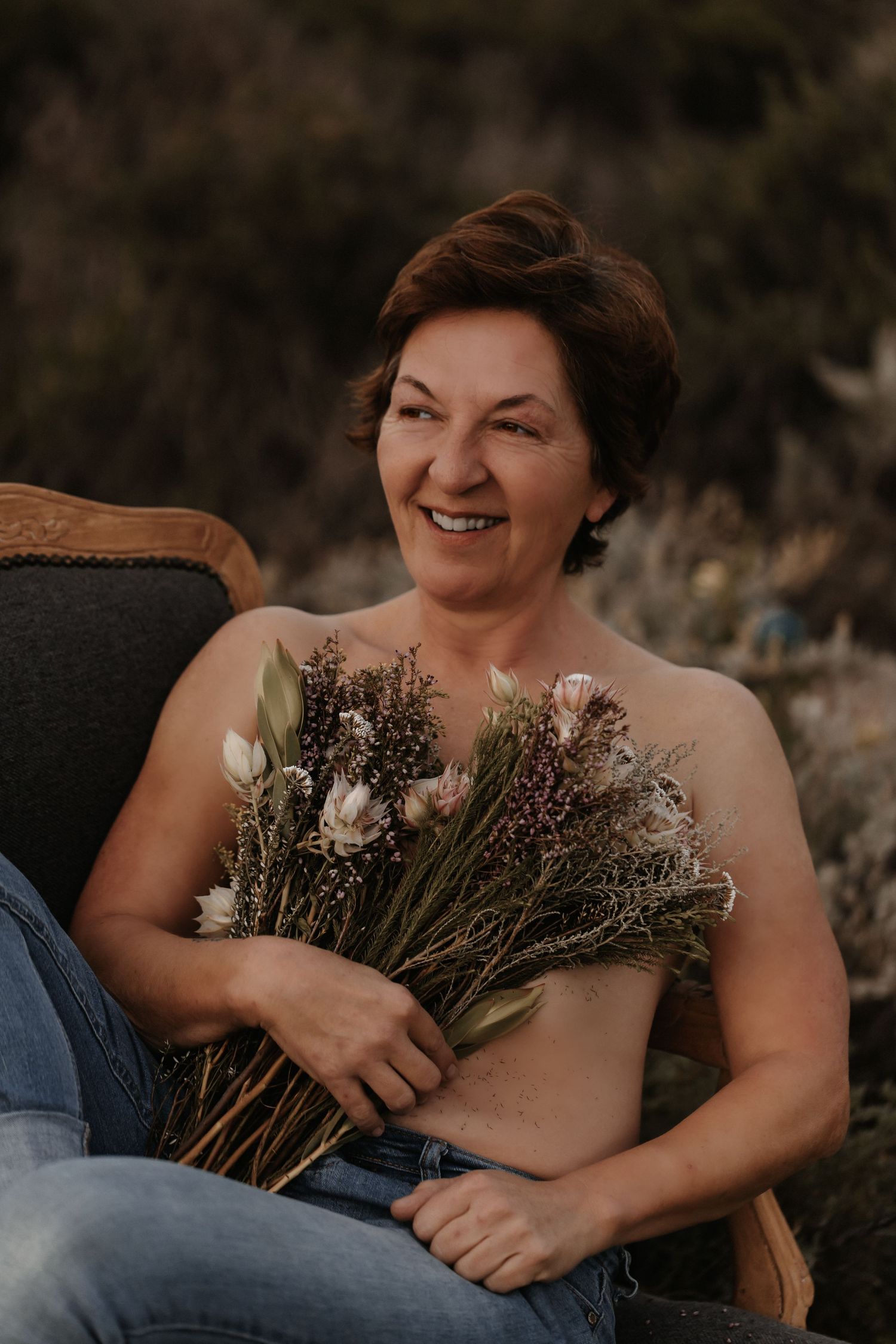 Person sits comfortably outdoors holding dried flower bouquet against natural backdrop in candid boudoir photoshoot.