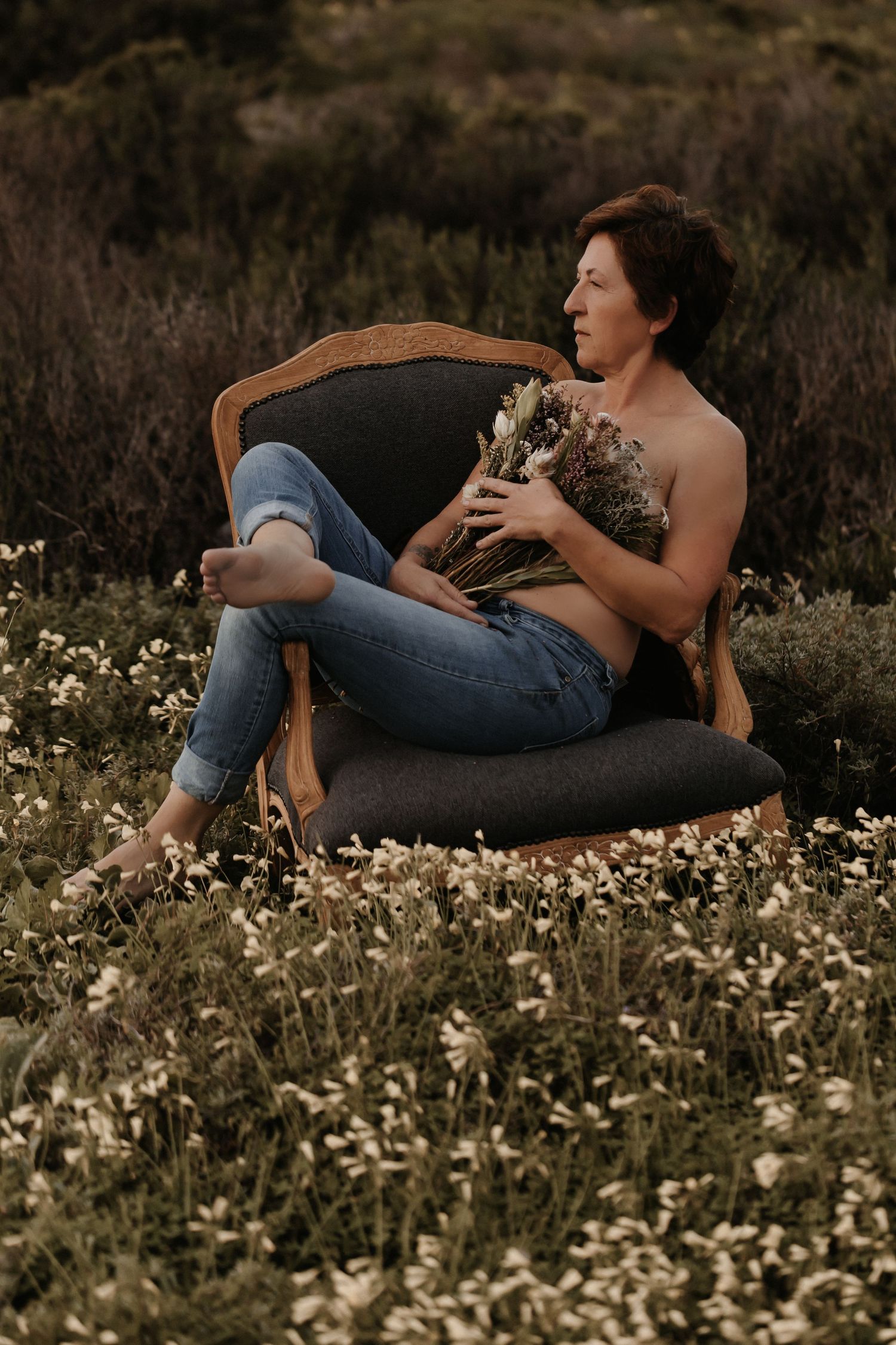 Person relaxing in a vintage armchair surrounded by wildflowers at sunset holding a small dog.