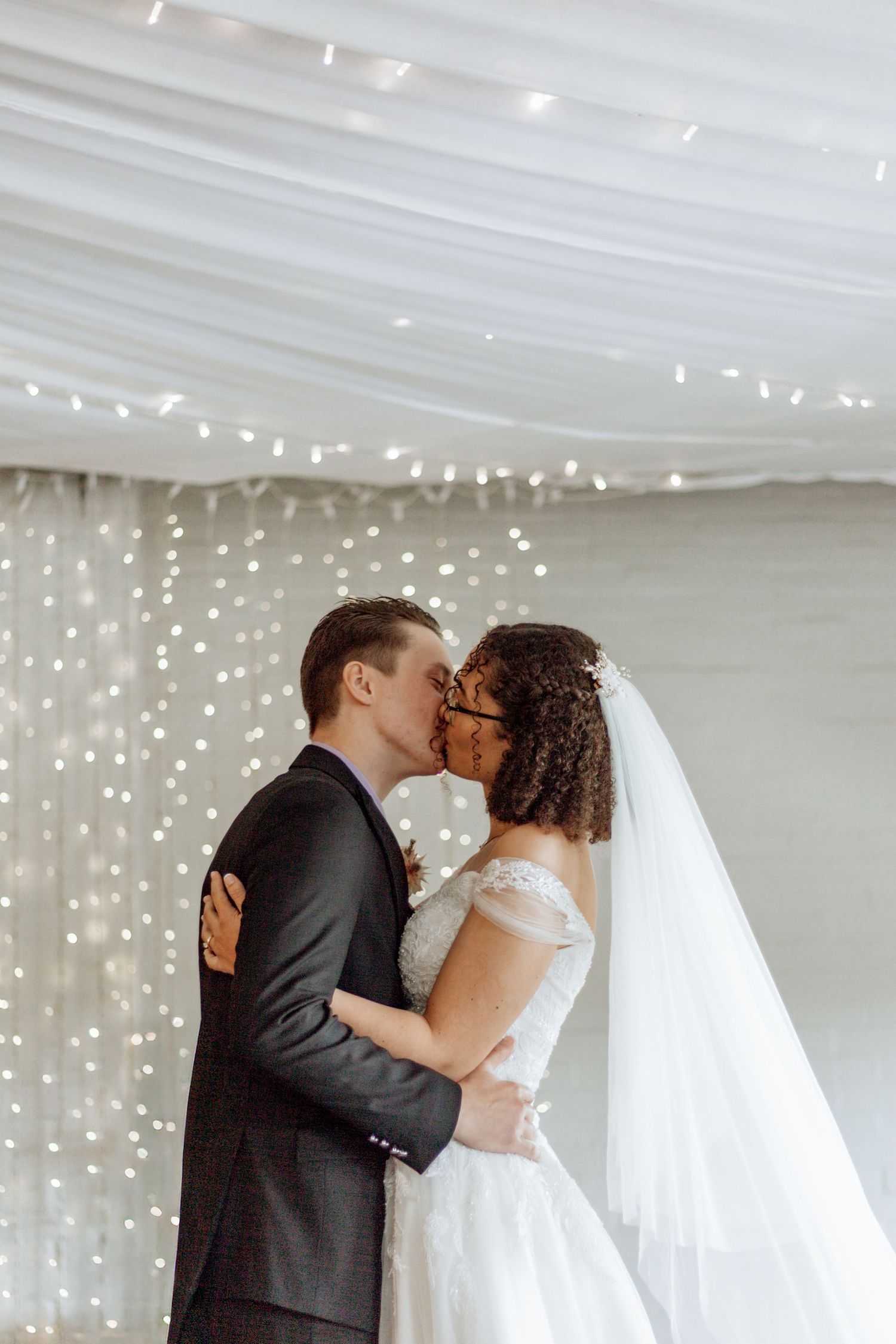 A romantic first kiss between newlyweds under twinkling fairy lights at an indoor wedding reception venue.
