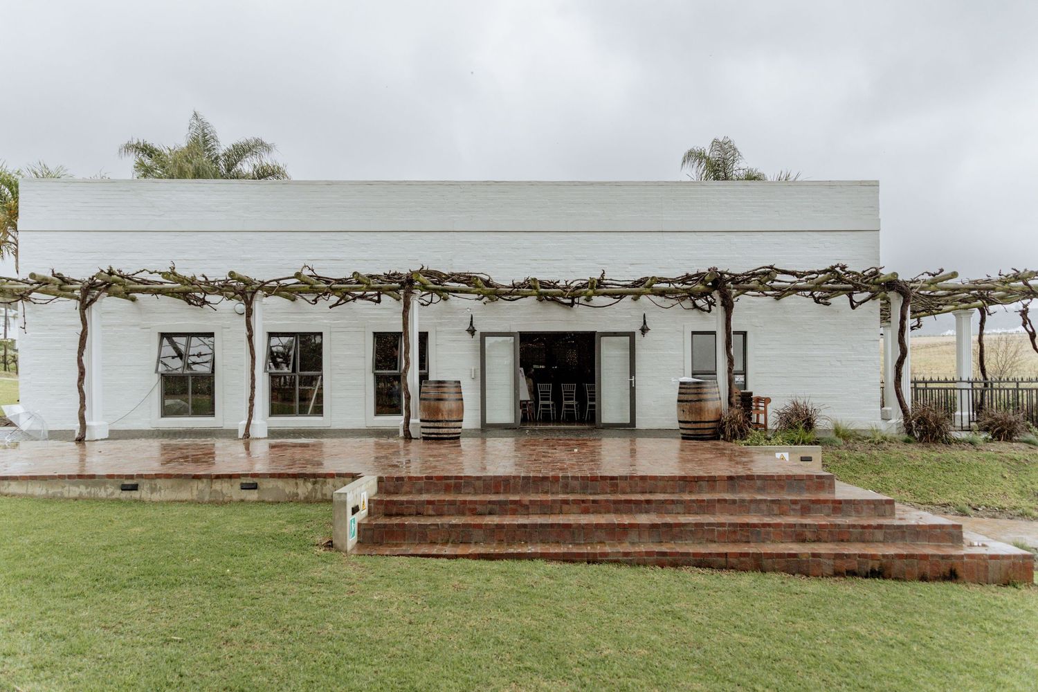 White stucco building with rustic wooden pergola and brick steps leading to covered patio area surrounded by green lawn.