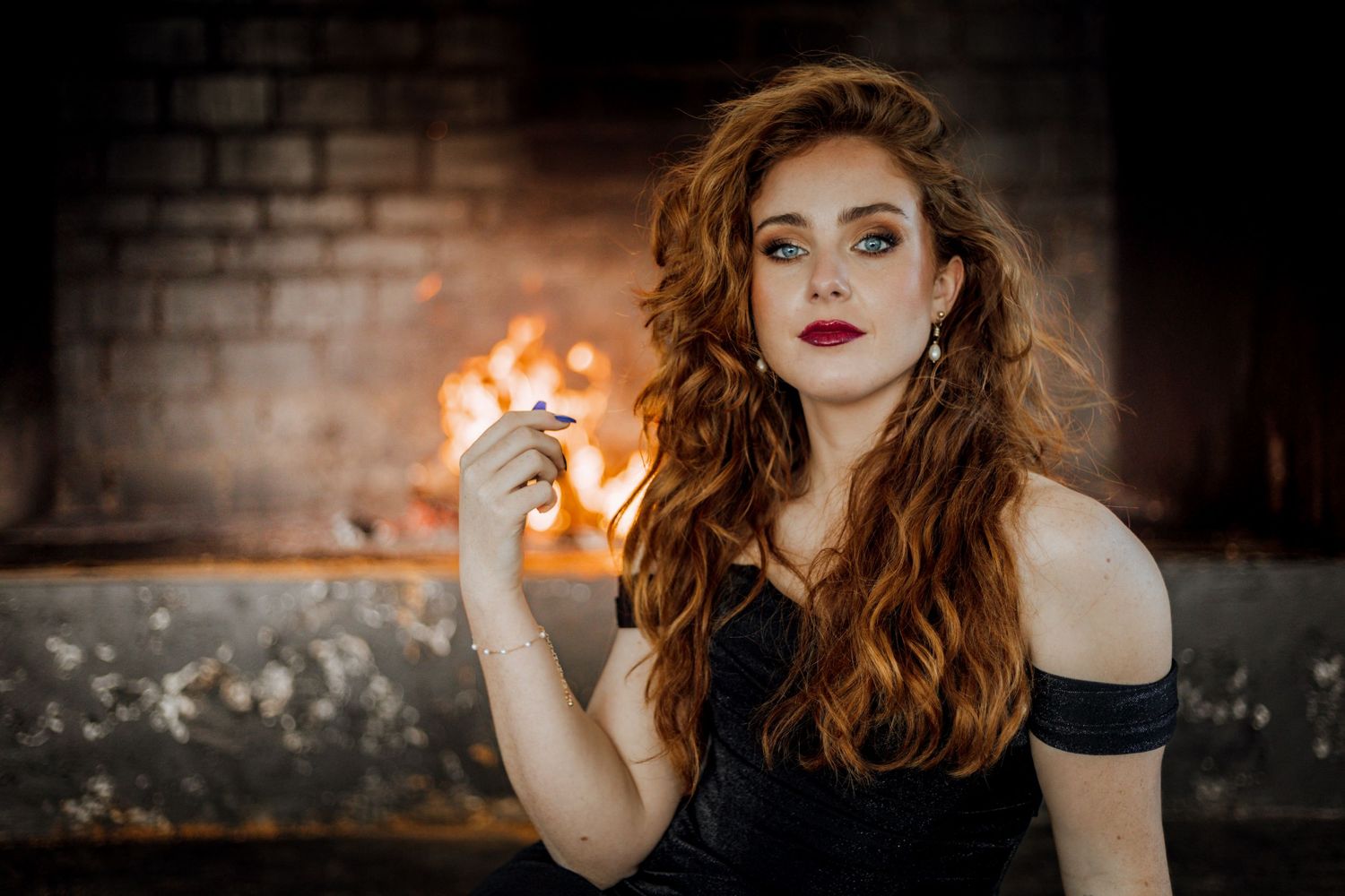 Dramatic profile portrait of woman with red hair and lipstick against dark brick wall with glowing flame.