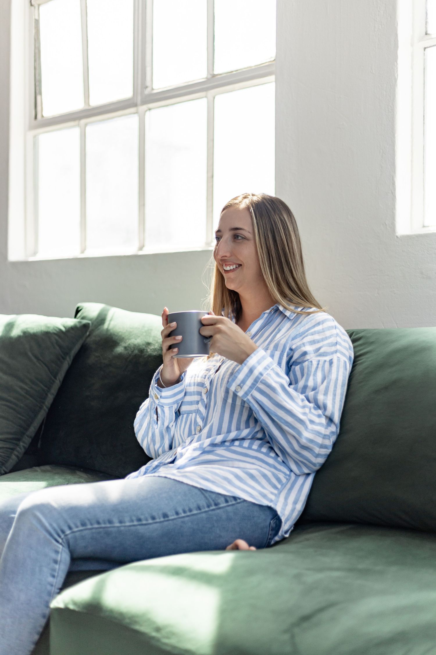 Person lounging comfortably on a dark green sofa while using a mobile phone in a sunlit room.
