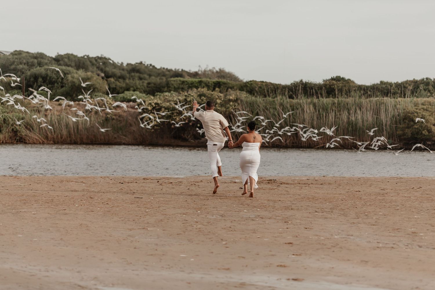 Two people running on a beach surrounded by soap bubbles at sunset with coastal vegetation in background.