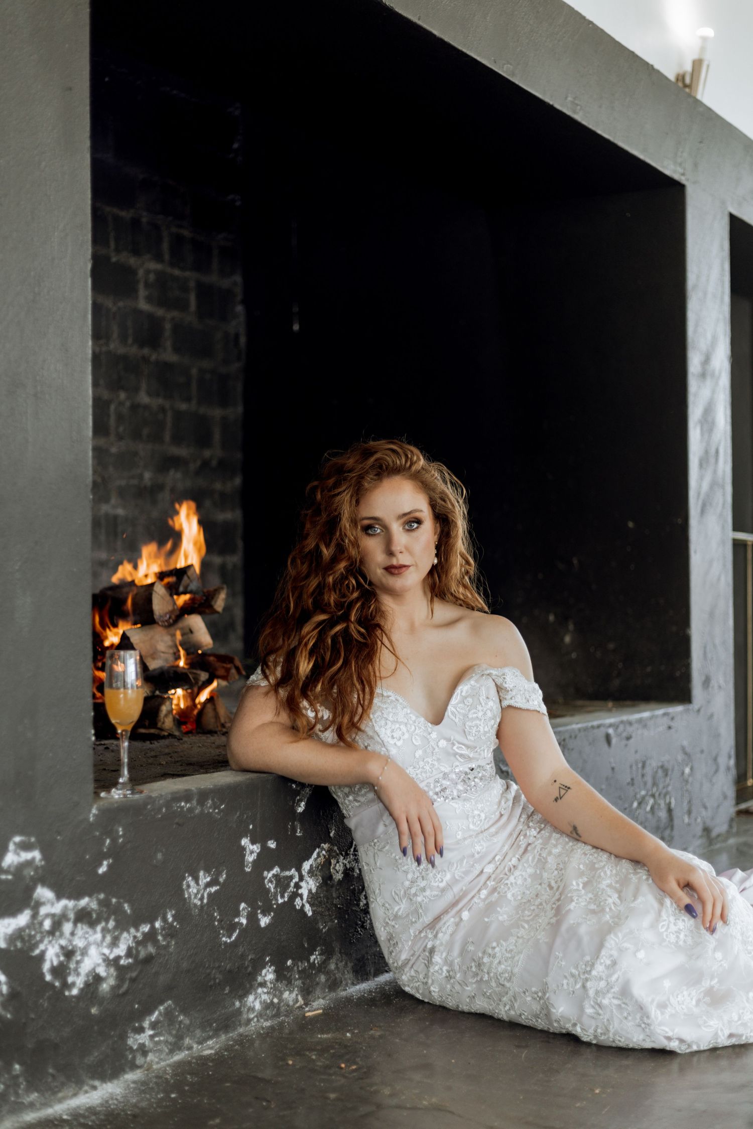 A bride in a detailed white lace wedding gown poses gracefully on the ground against a rustic wall with a fireplace.