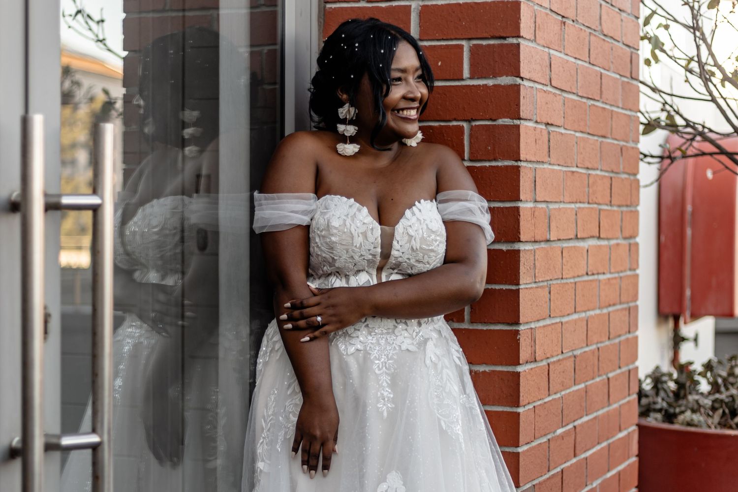 A bride in a white lace wedding gown poses against a red brick wall and window backdrop.