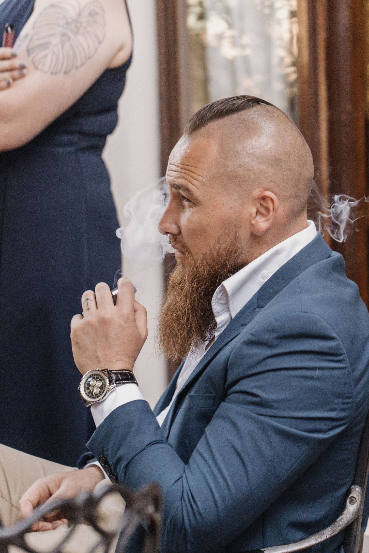 A man in a blue suit with a long beard exhales smoke while wearing a luxury watch during a formal event.
