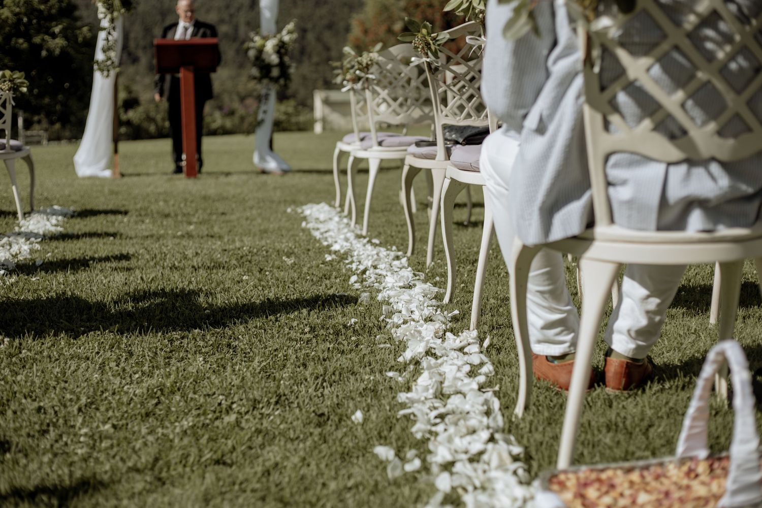 White chairs line an outdoor wedding aisle decorated with delicate white floral petals and greenery on a sunny day.