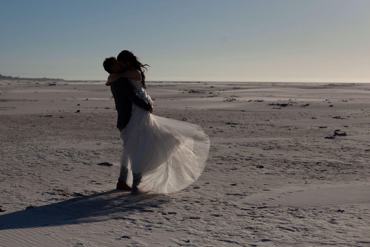 Couple embracing on a sandy beach, bride in flowing white gown and groom in dark suit, with clear blue sky backdrop.