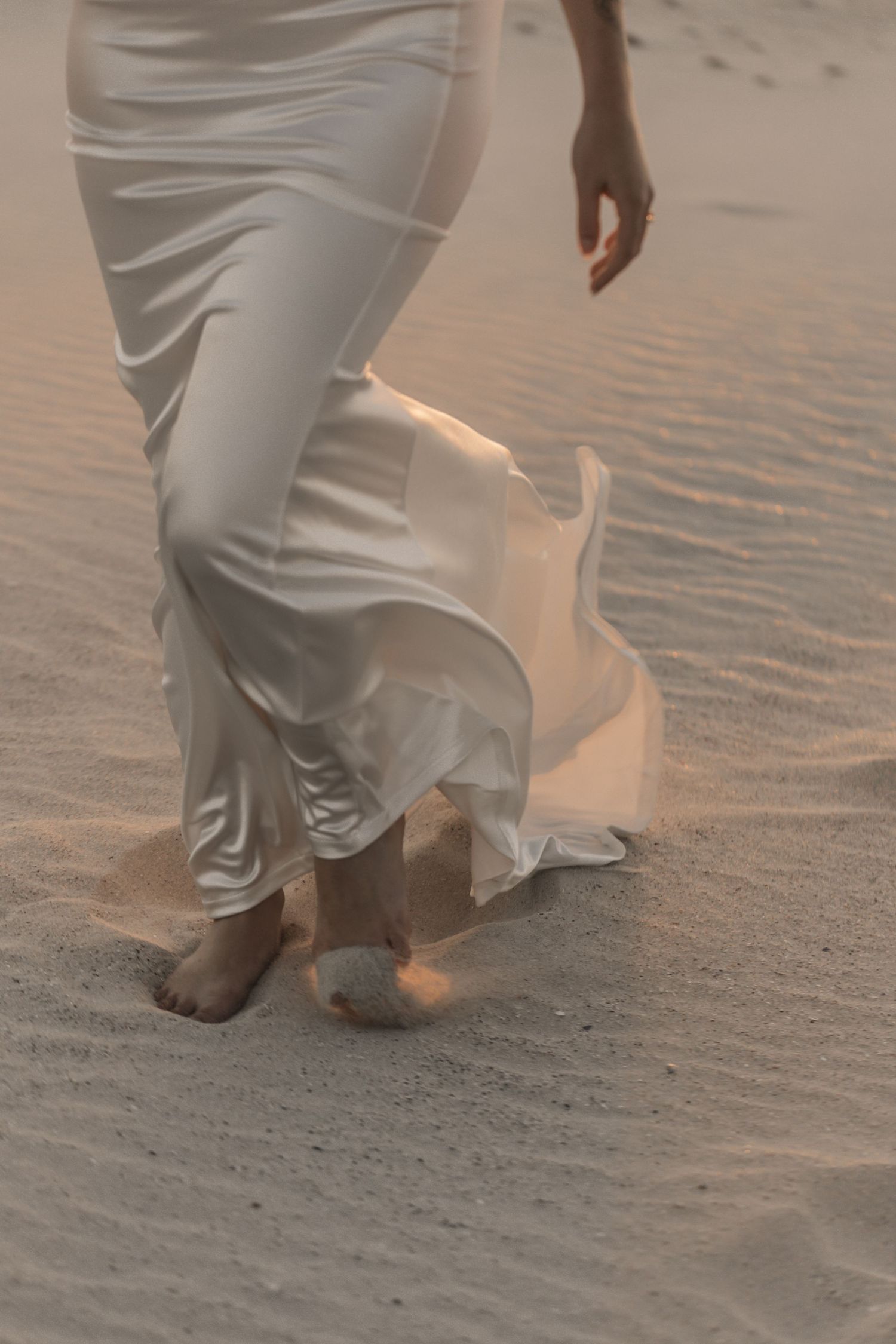 Barefoot bride walking on sand dunes in flowing white satin dress, fabric billowing in the wind.