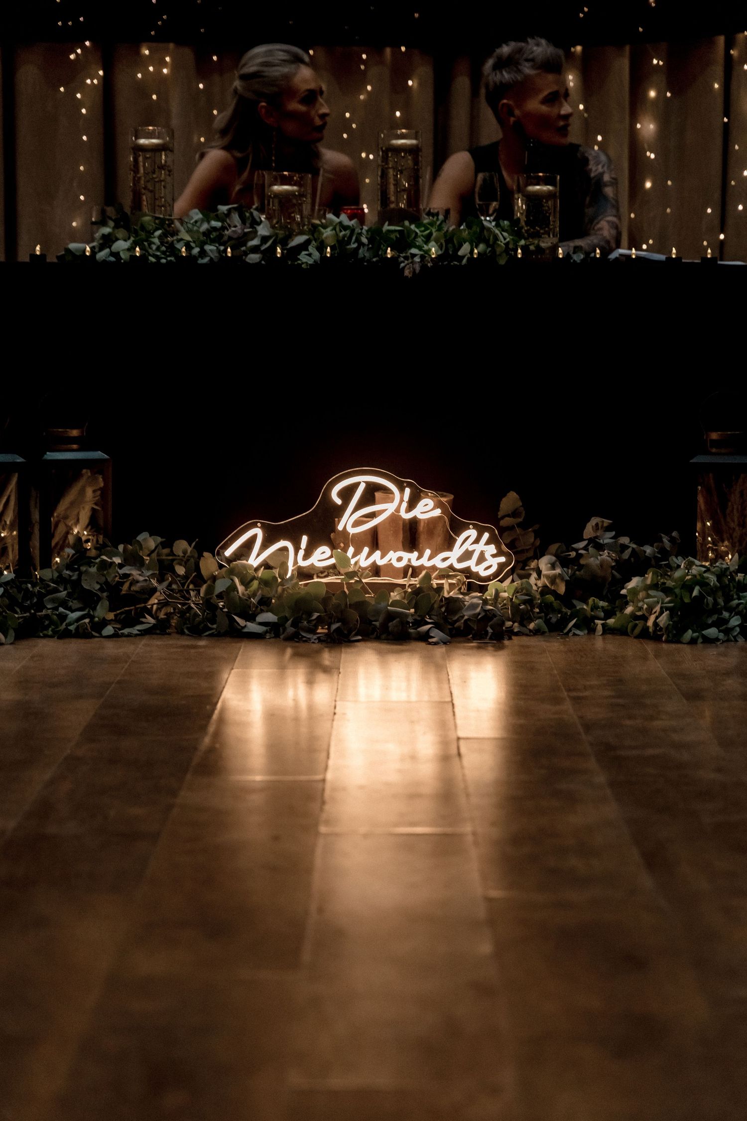 A glowing neon sign reading 'The Morning After' illuminates a dark room with greenery and string lights in the background.