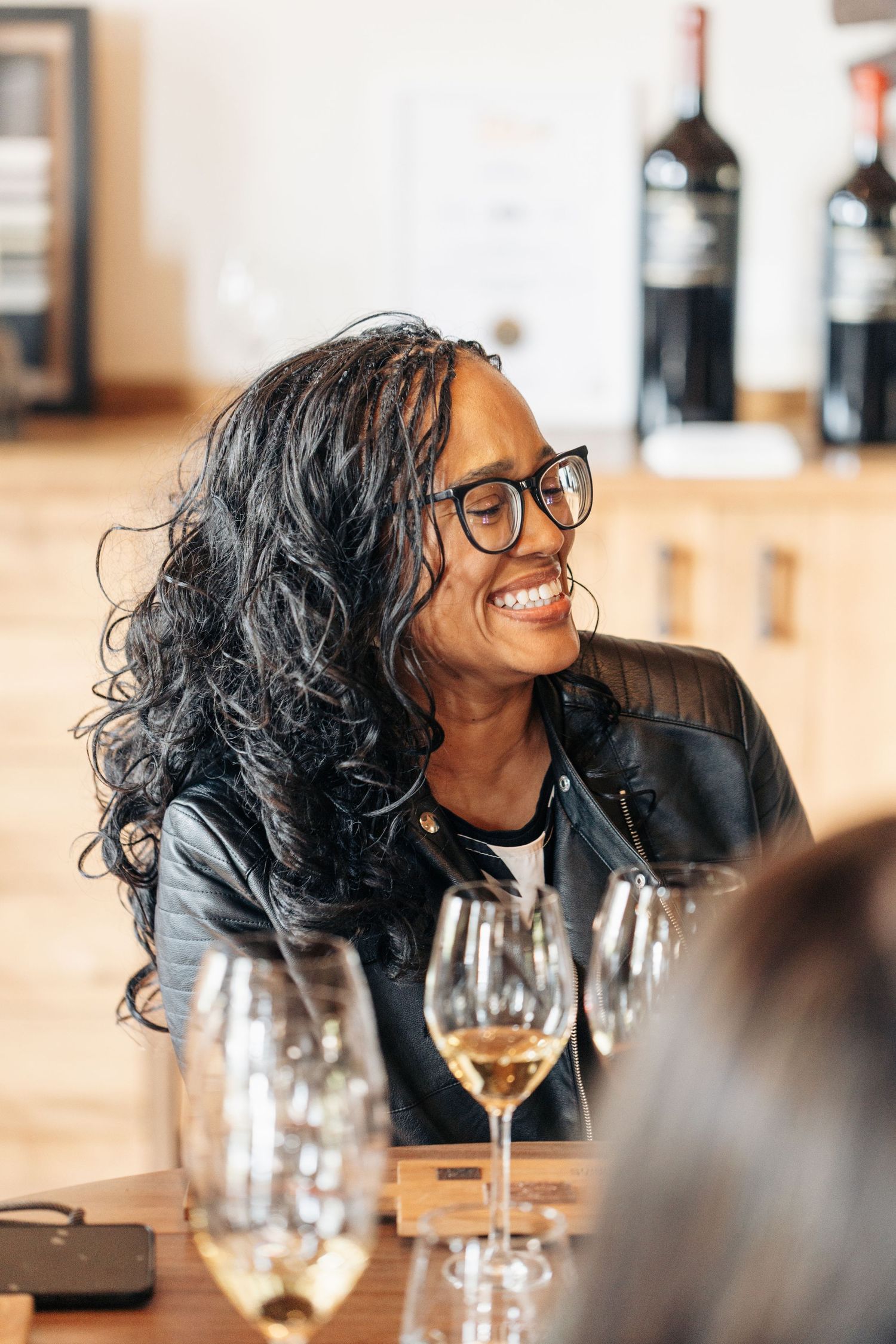 A smiling woman with glasses enjoys a wine tasting at a winery, with glasses of white wine on the table.