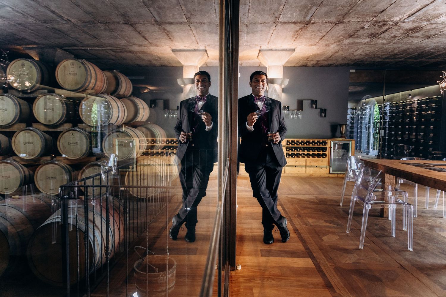 Man in black suit reflected in mirror surrounded by wine barrels and bottle storage in elegant winery cellar.