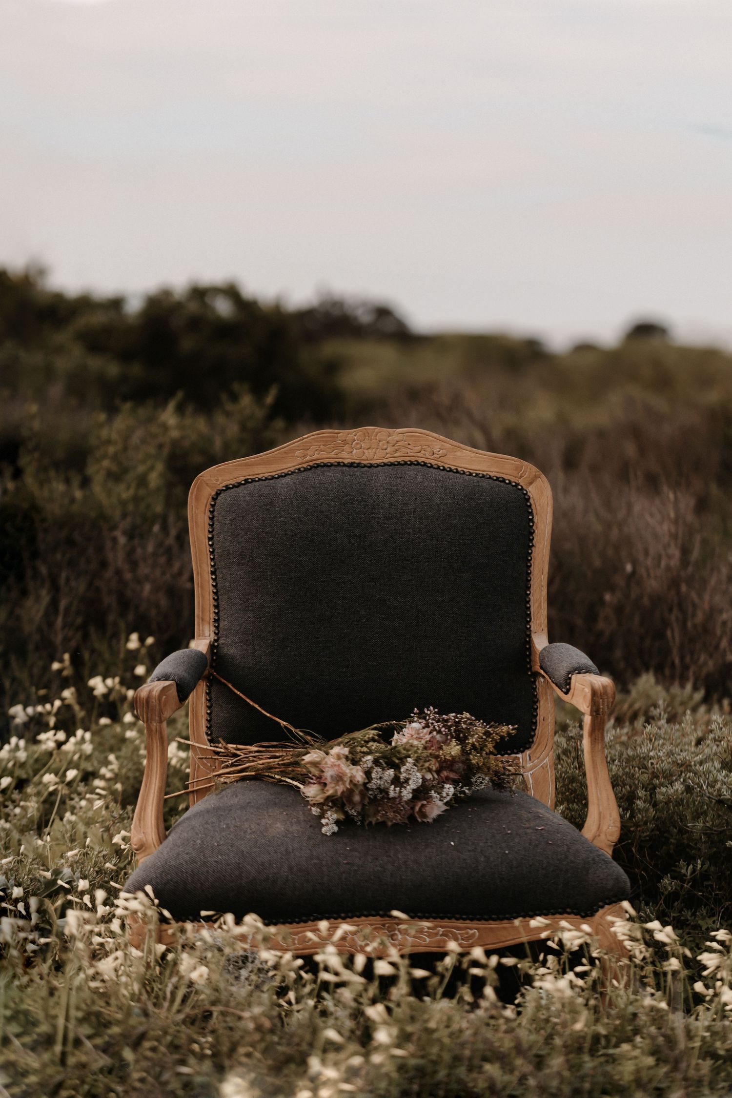 A vintage charcoal gray armchair with wooden frame sits in a meadow with dried flowers placed on the seat.