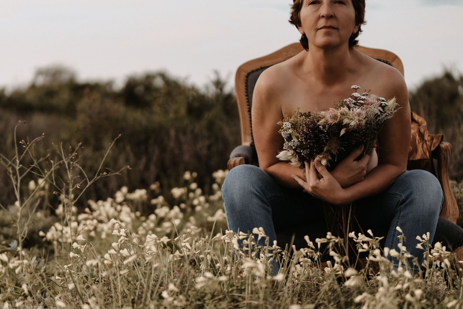 Someone sits cross-legged in a wildflower field at sunset holding a rustic bouquet while wearing jeans and strapless top.