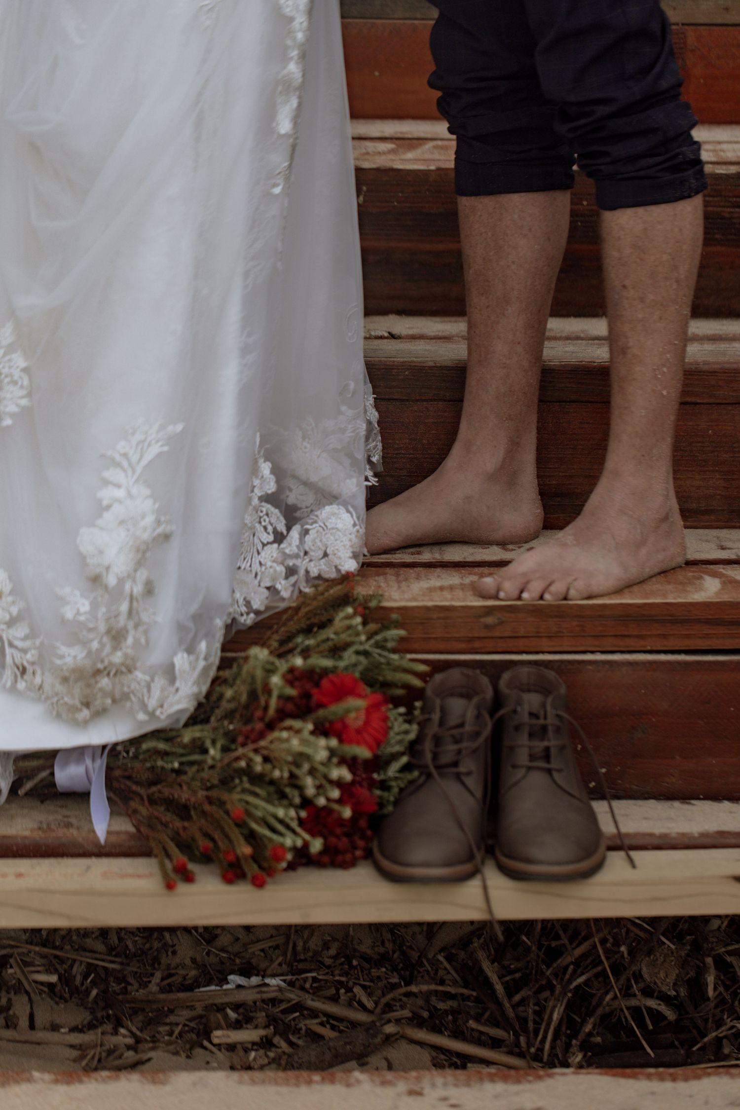 Wedding details with bare feet on wooden steps next to a white lace dress, shoes, and red bouquet on rustic stairs.