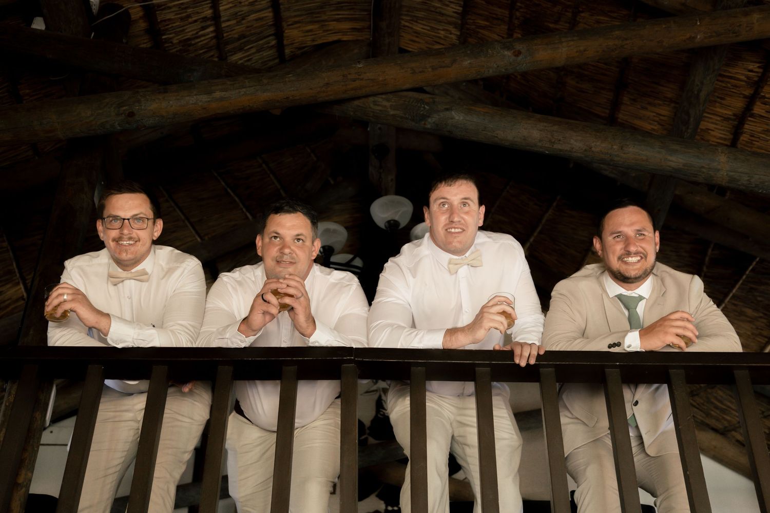 Four groomsmen in tan suits lean on a wooden balcony railing, smiling at the camera in a rustic venue.