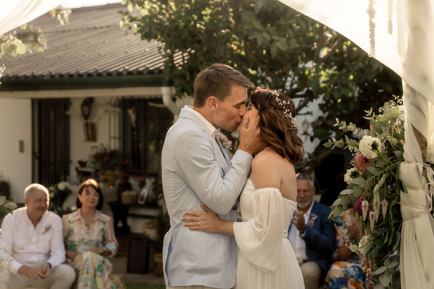 Couple shares first kiss at outdoor wedding ceremony with guests seated behind them and floral wreath decoration.