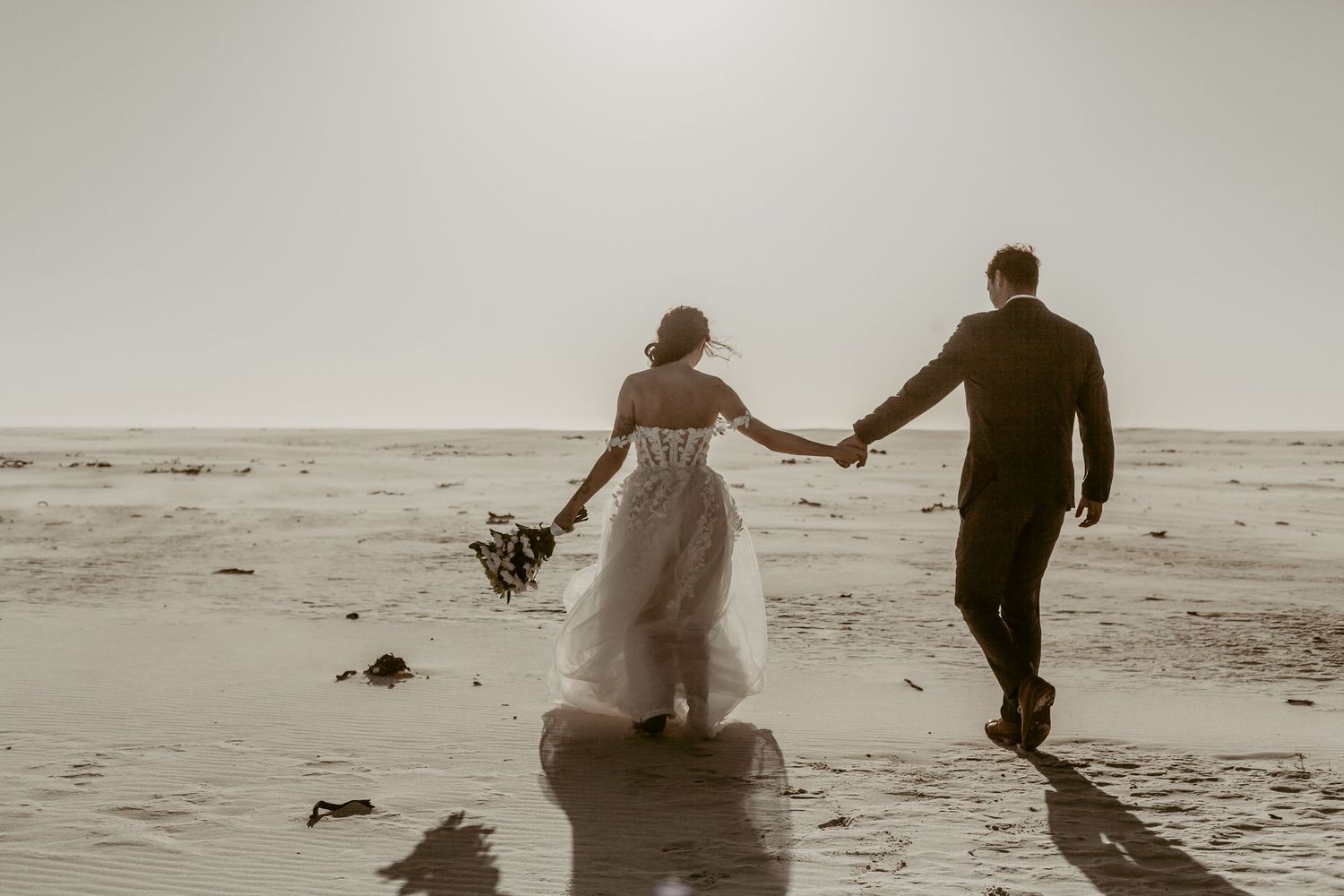 Bride and groom holding hands walking toward ocean on beach in wedding attire with bouquet in golden sunlight.
