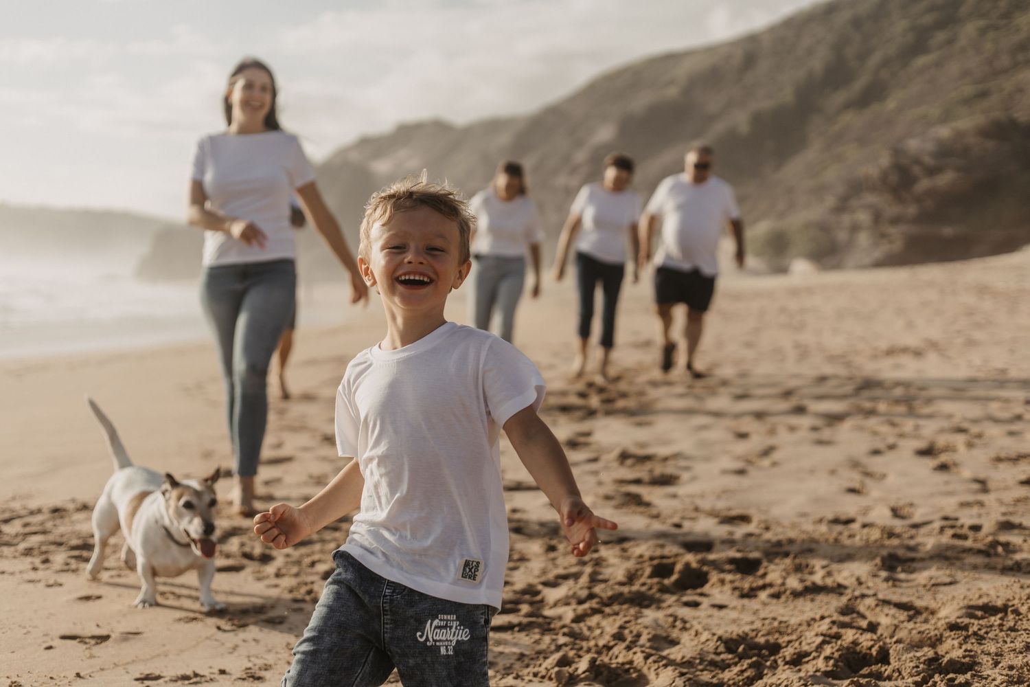 A joyful child runs along a sandy beach with family members and a small dog following behind on a misty day.