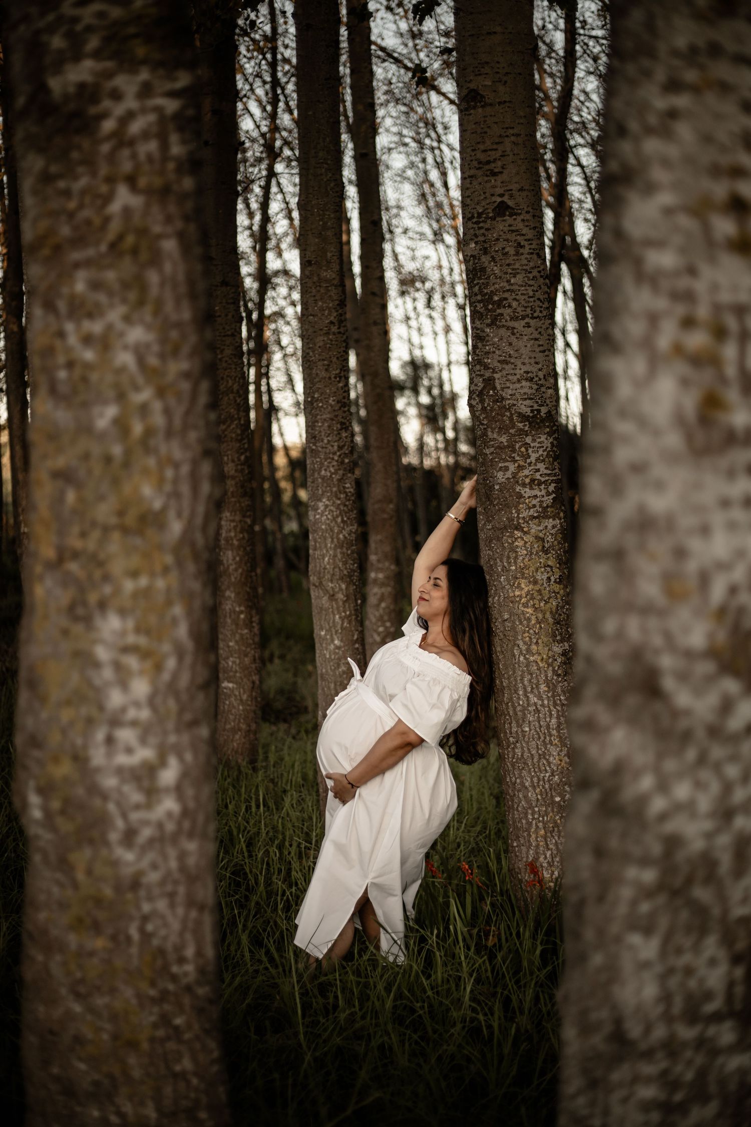 A woman in a flowing white dress poses among tall trees in a dreamy forest maternity photoshoot at sunset.