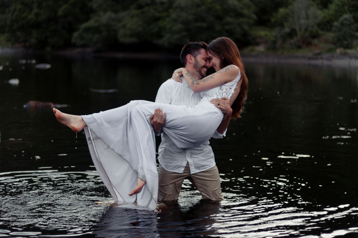 A couple in white clothing dances together in a lake during a romantic photoshoot at sunset.