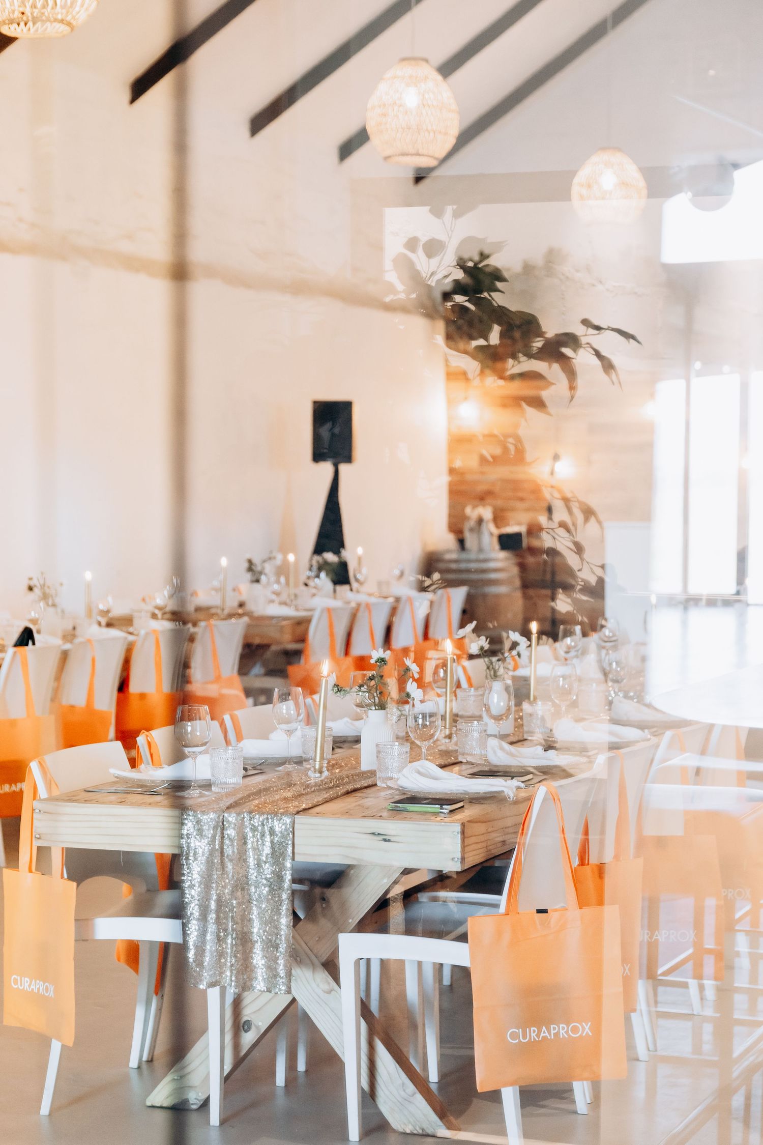 Elegant event venue with white chairs, wooden tables, sequin runners, and orange CuraBox gift bags in a bright, airy space.