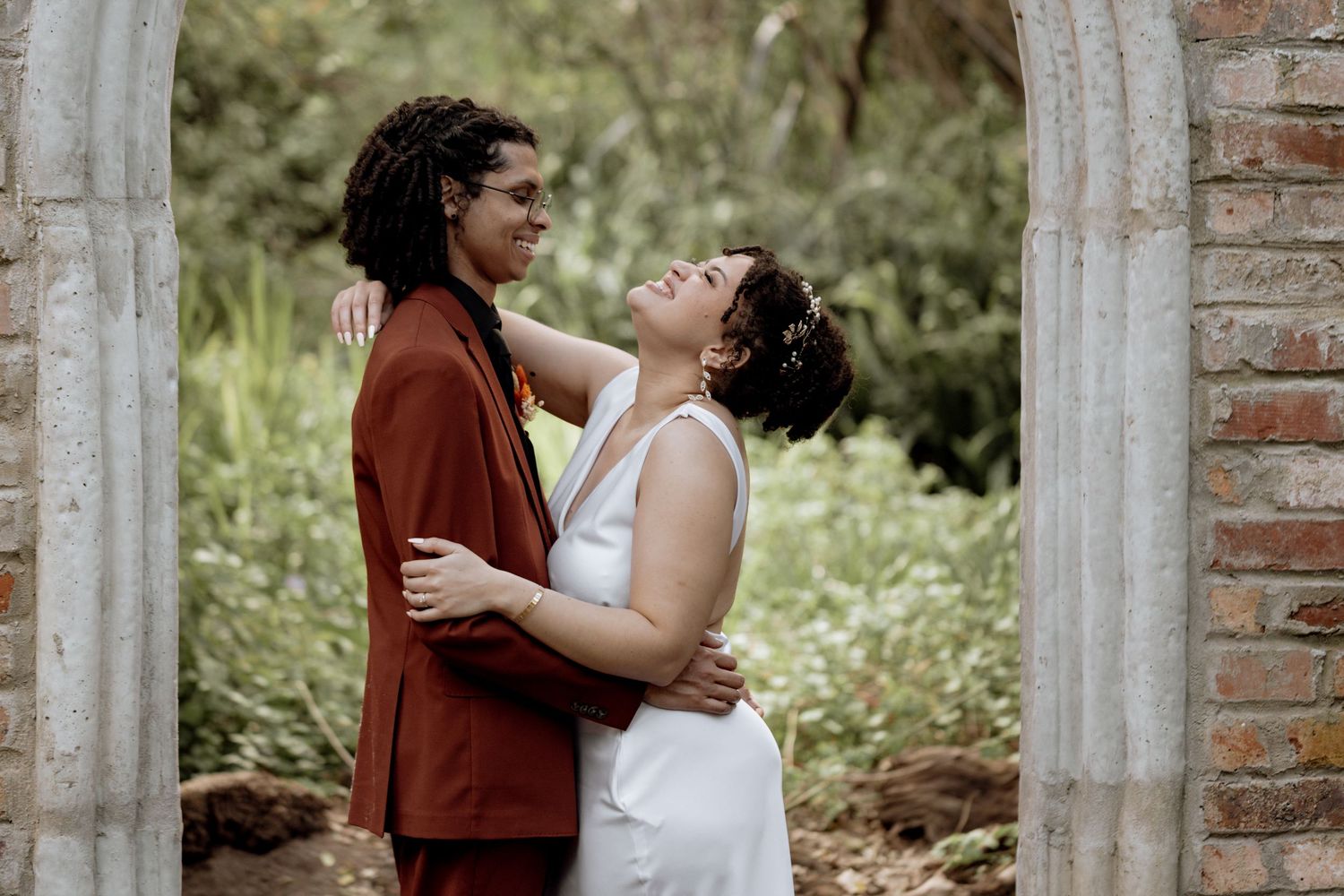 A couple in wedding attire share an intimate moment between two stone pillars in a garden setting.