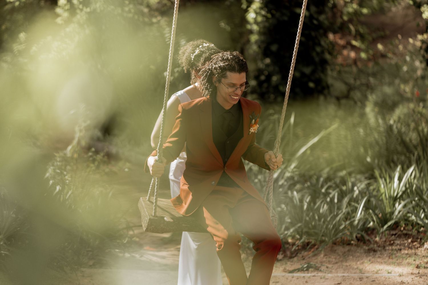 A person sits on a swing in a sunlit garden, wearing brown and white casual attire while looking downward.