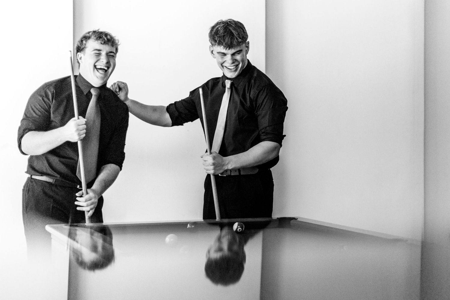 Two smiling men holding pool cues lean over a billiard table in a black and white photo.