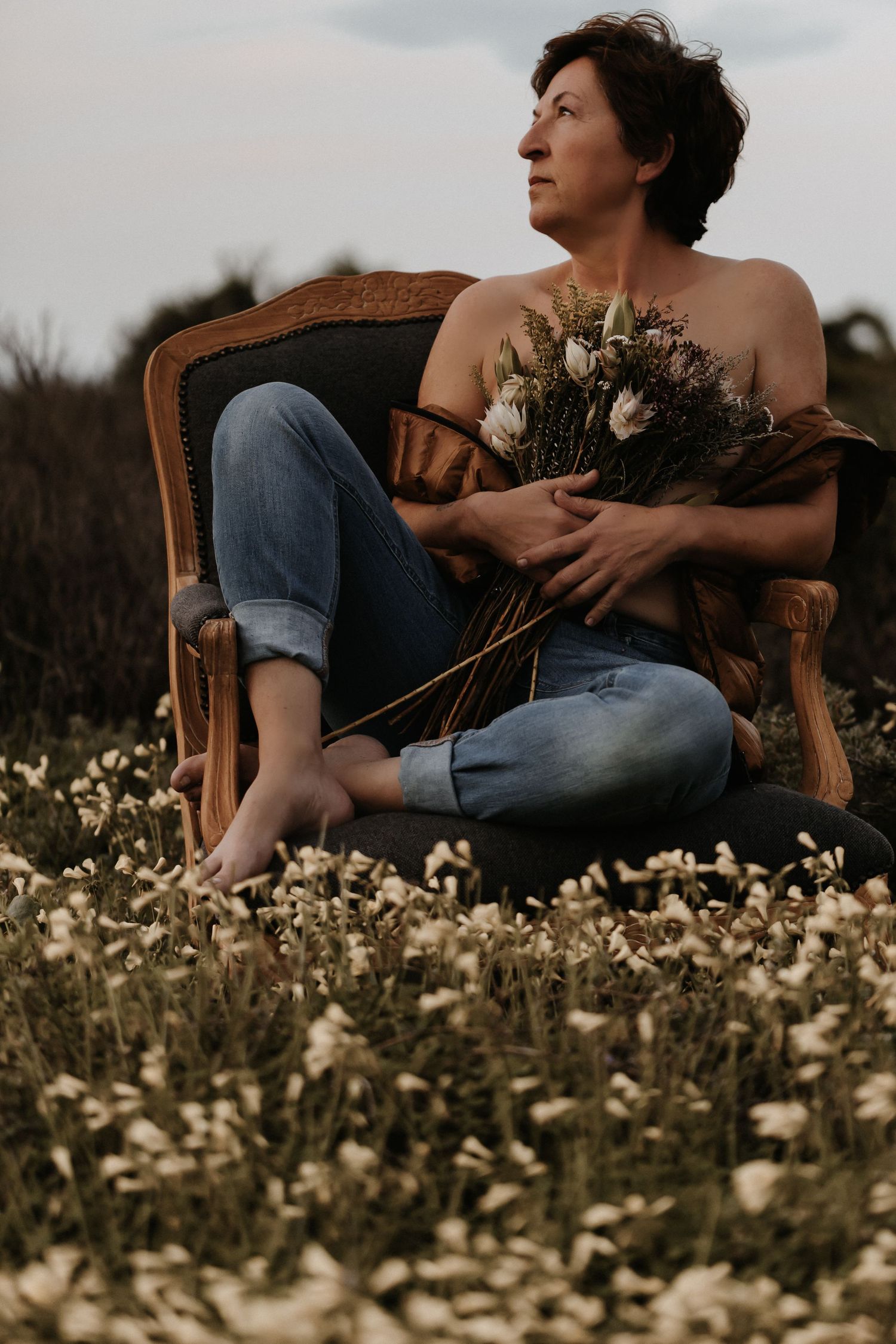 A moody portrait in a vintage chair surrounded by wildflowers shows someone holding dried flowers at sunset.