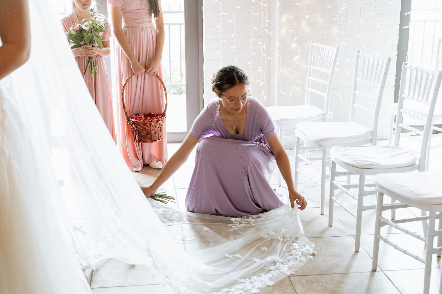 Multiple bridesmaids dressed in pink gather around a white dress, helping with wedding preparations in a light filled room.