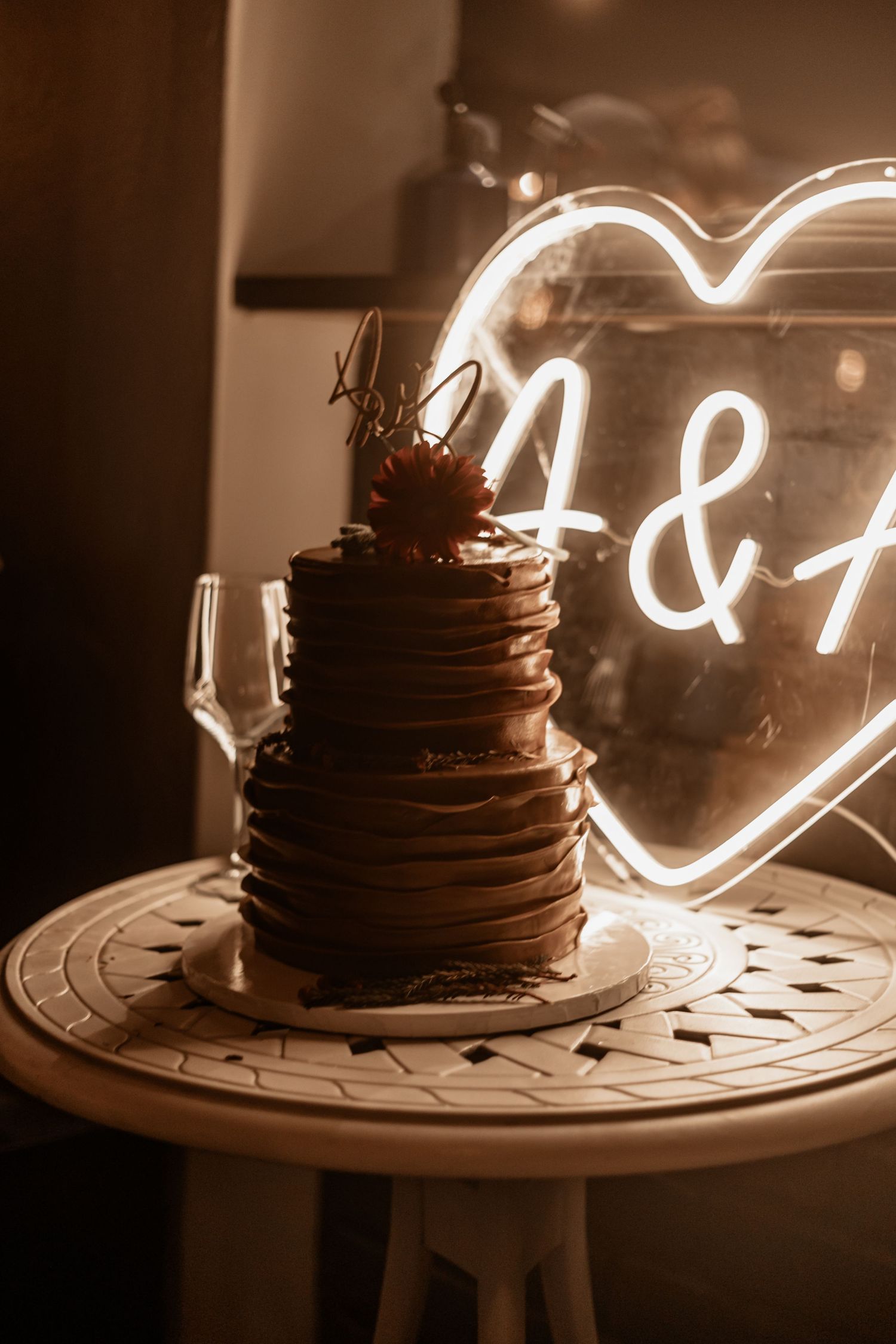 Chocolate wedding cake displayed on rustic table with glowing neon heart sign in warm sepia tones.
