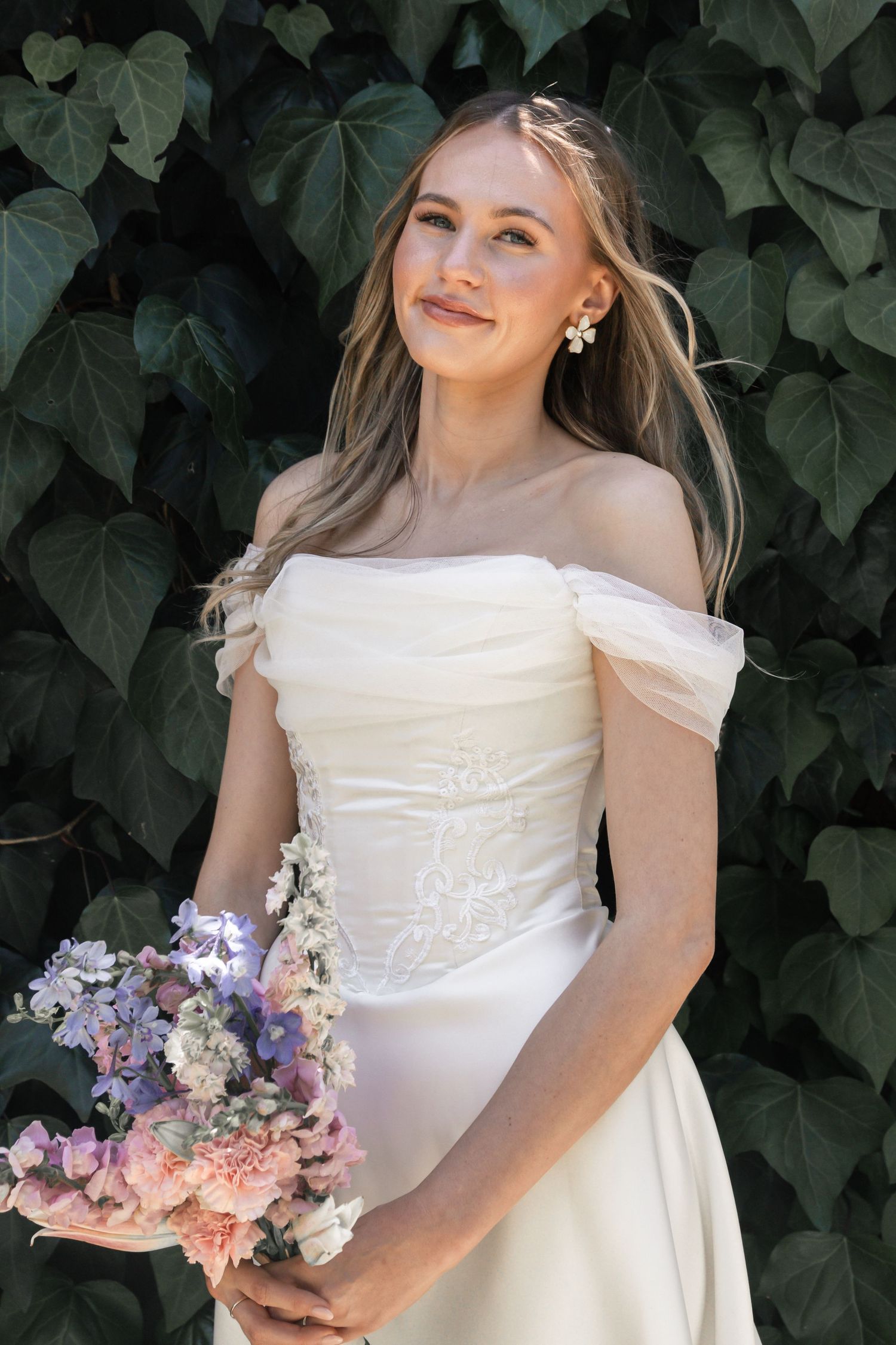 A bride in an elegant white gown holds a pastel bouquet against a backdrop of lush green foliage.