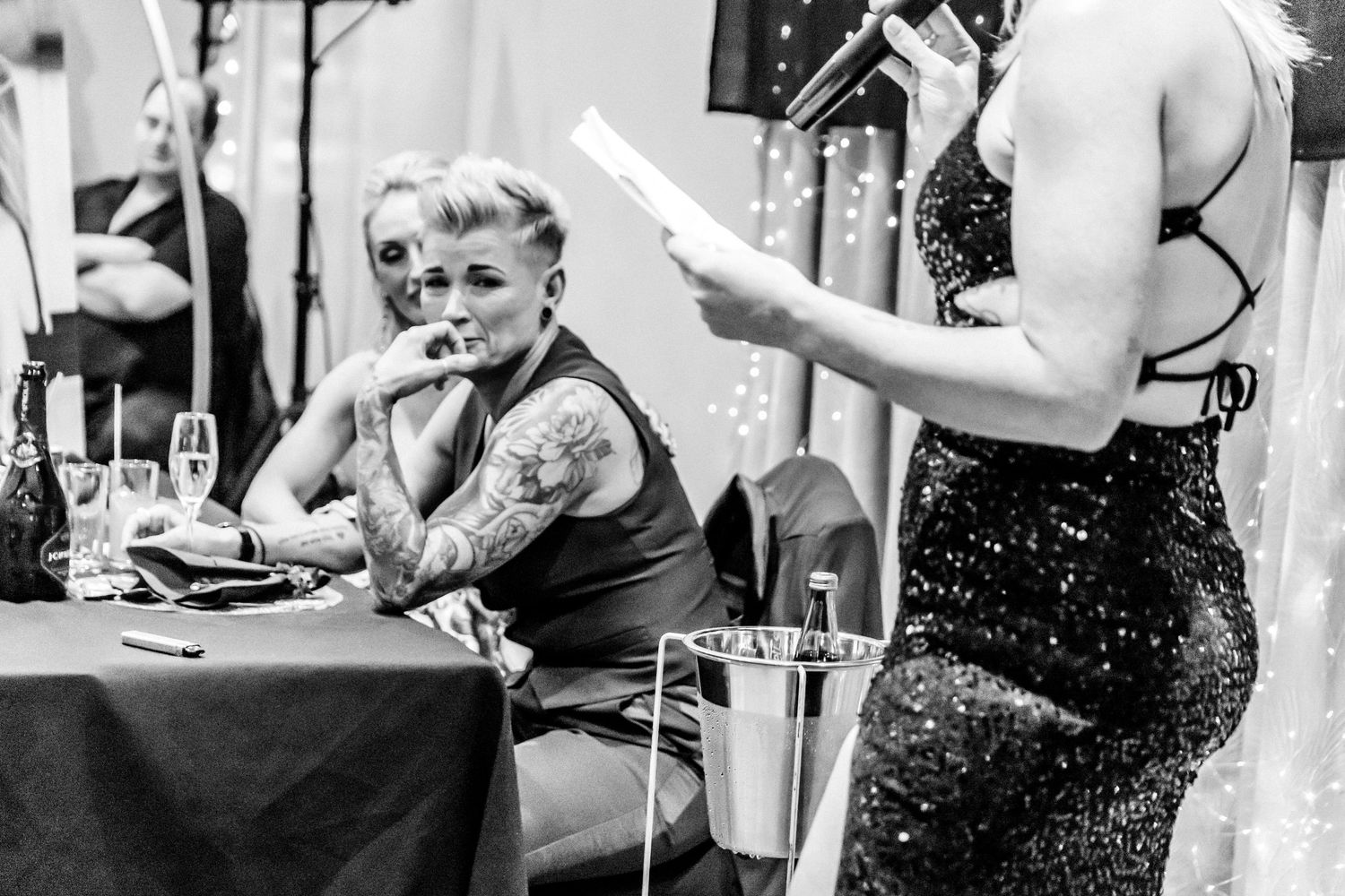 A black and white photo of a tattooed person sitting at a table while someone in a sequined dress reads from a paper.