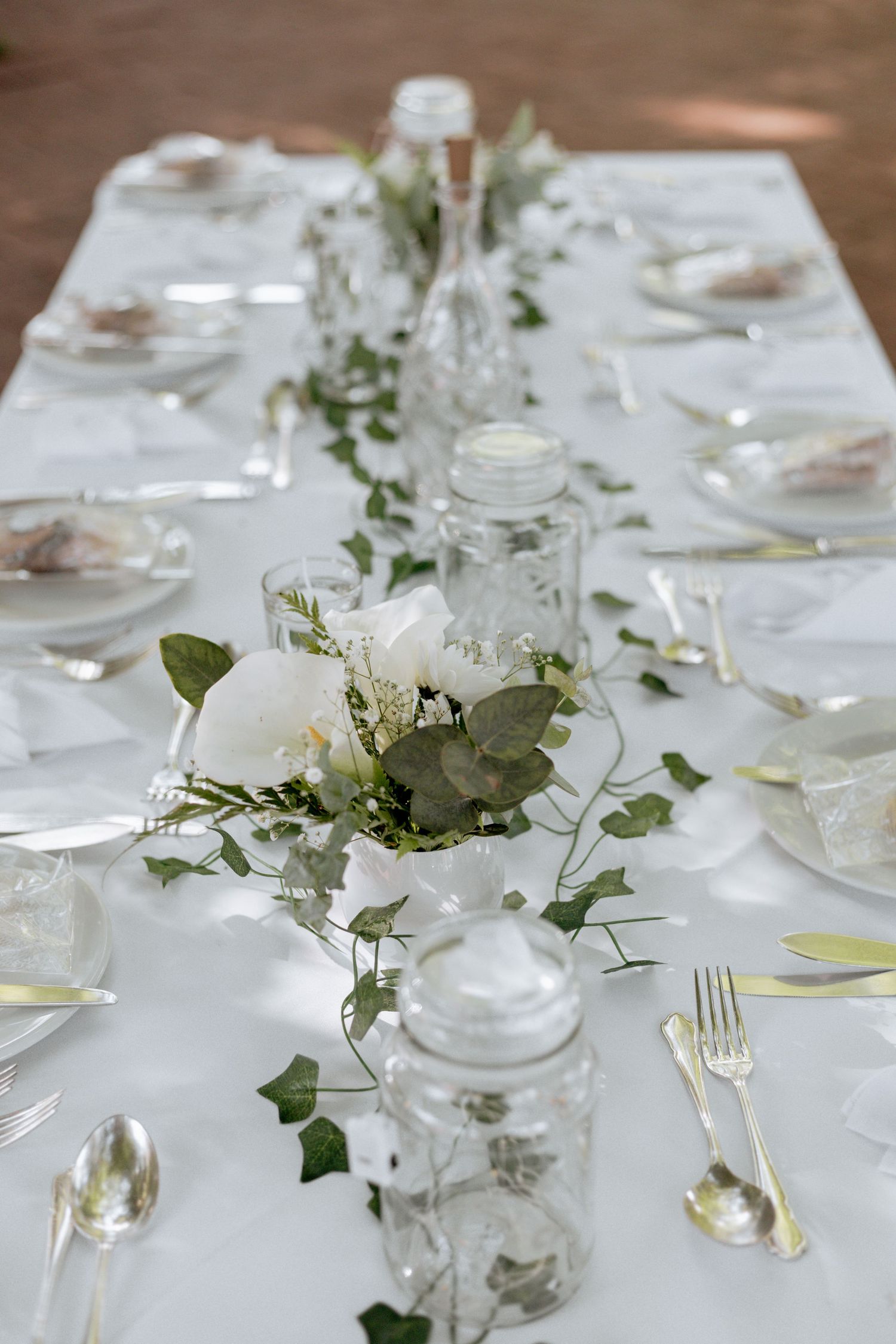 Elegant white tablescape with mason jars, white flowers, trailing greenery and vintage place settings on linen tablecloth.