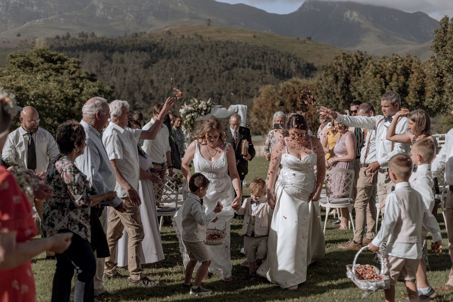 Wedding guests in white attire gather outdoors with scenic mountain views and greenery in the background.