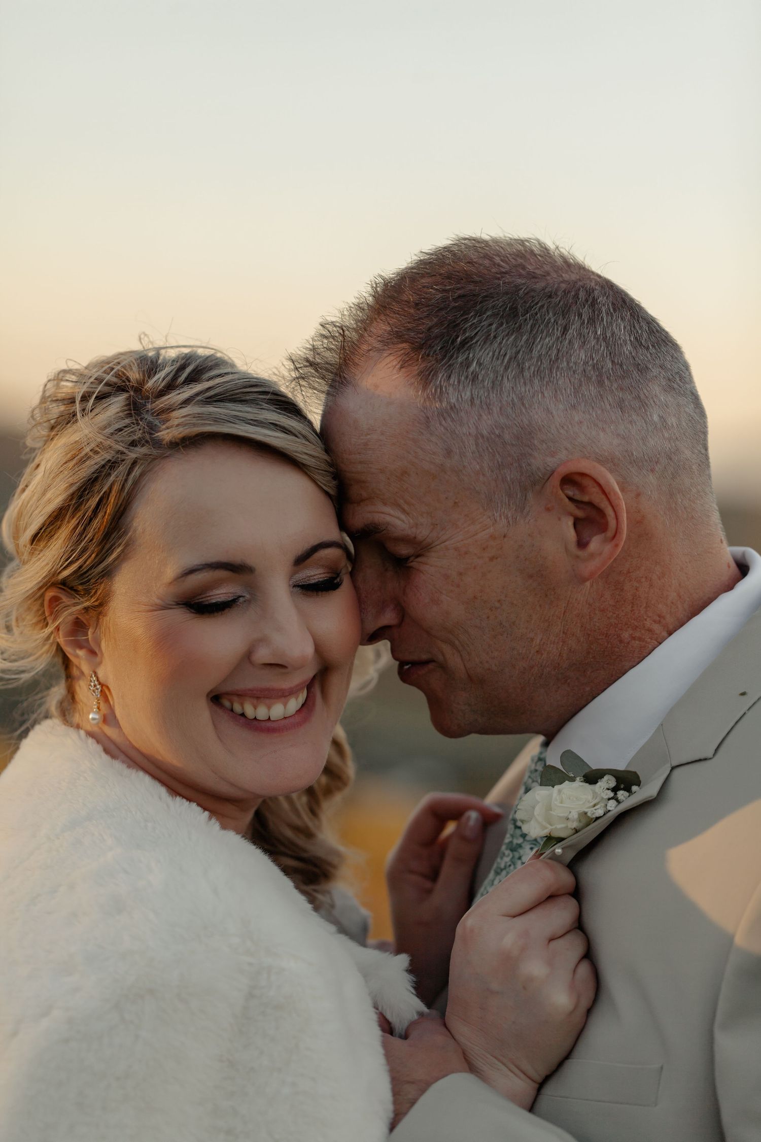 A joyful newlywed couple embraces in soft golden light, with the bride wearing a white fur wrap and groom in beige suit.