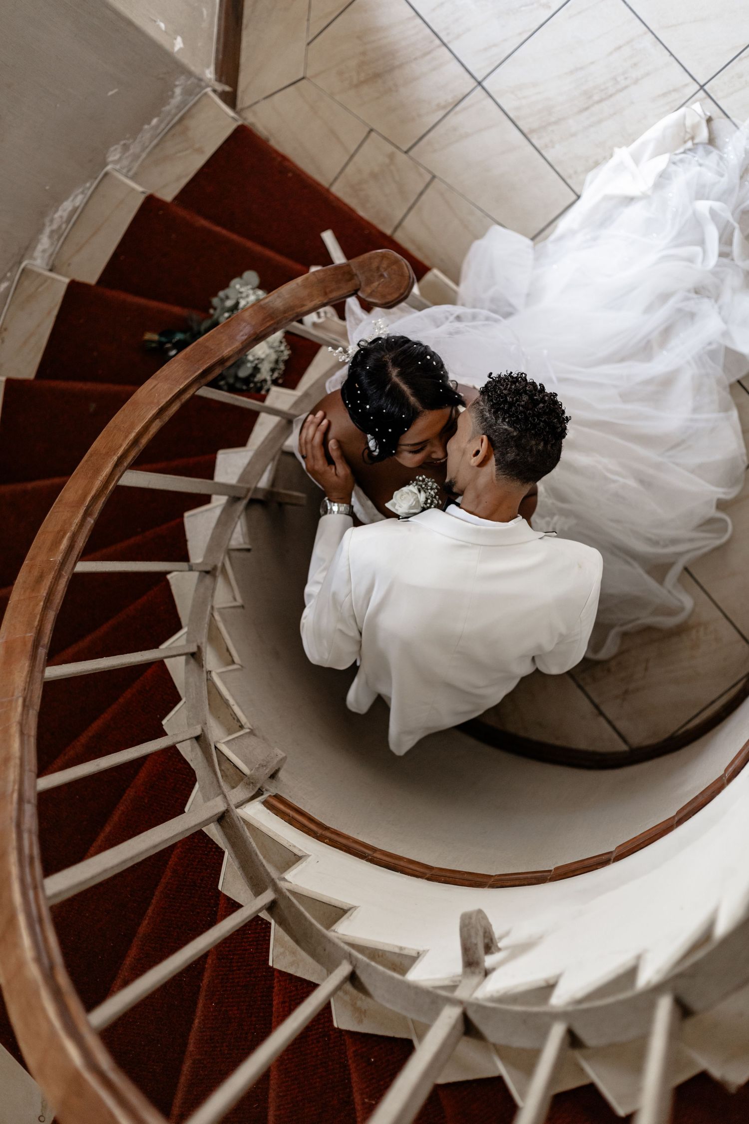 Overhead view of couple in wedding attire embracing on a spiral staircase with red carpet.