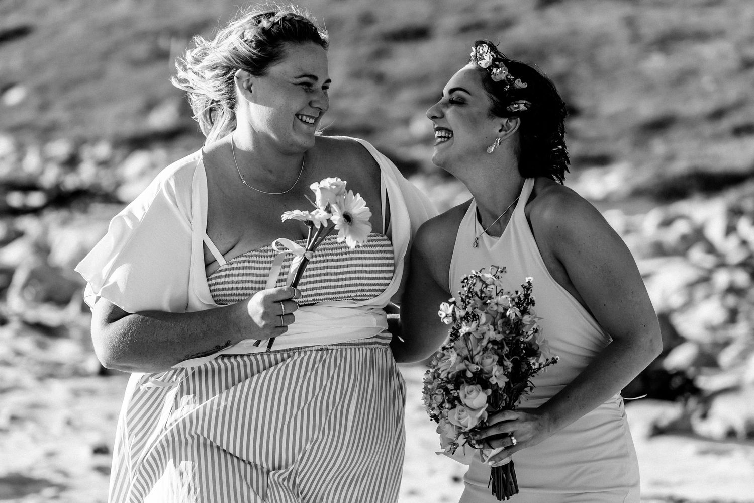 Two happy people in light dresses share a joyful moment at an outdoor setting while holding flowers.