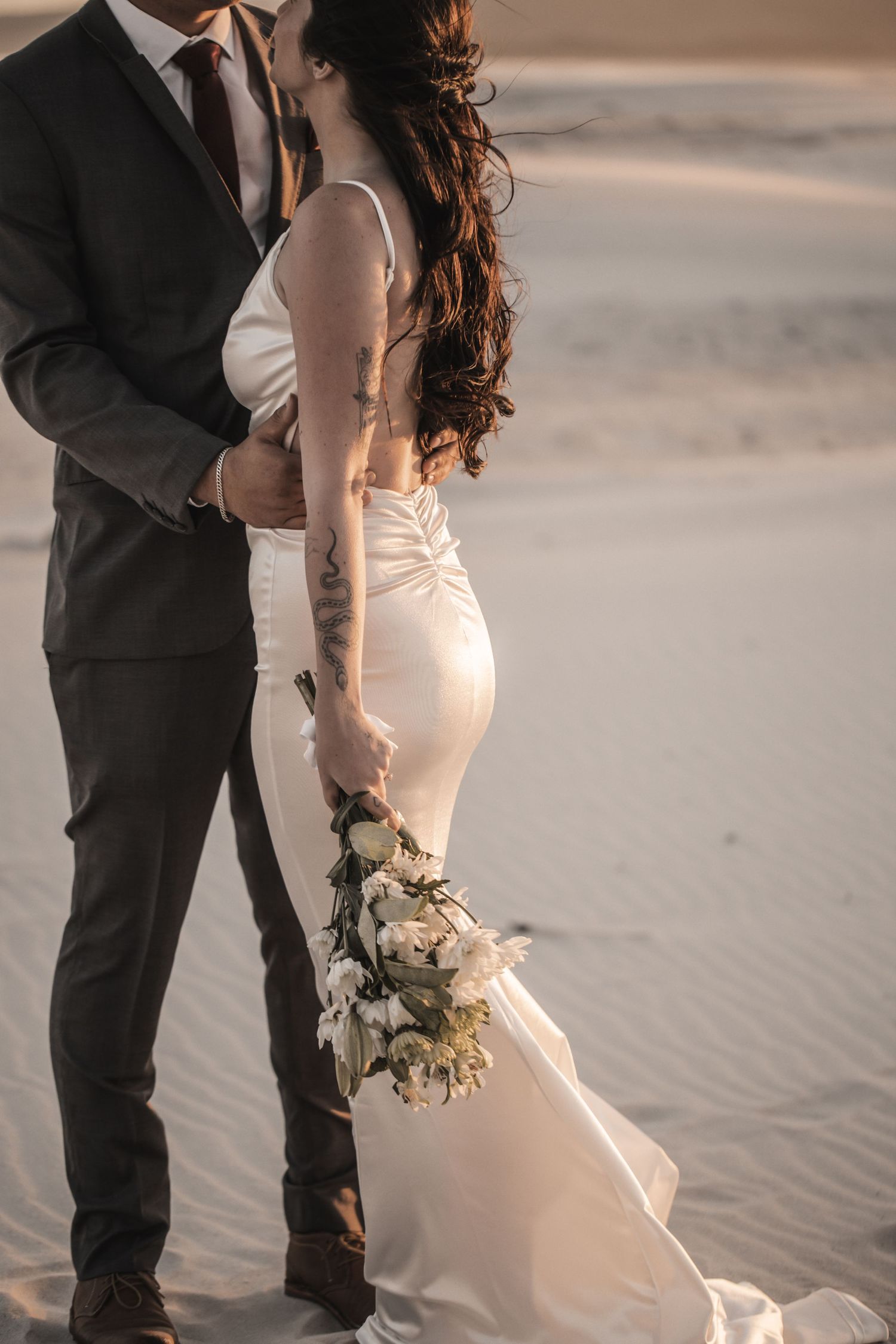 Newlywed couple embraces on sandy beach at sunset with bride in white dress holding delicate floral bouquet.