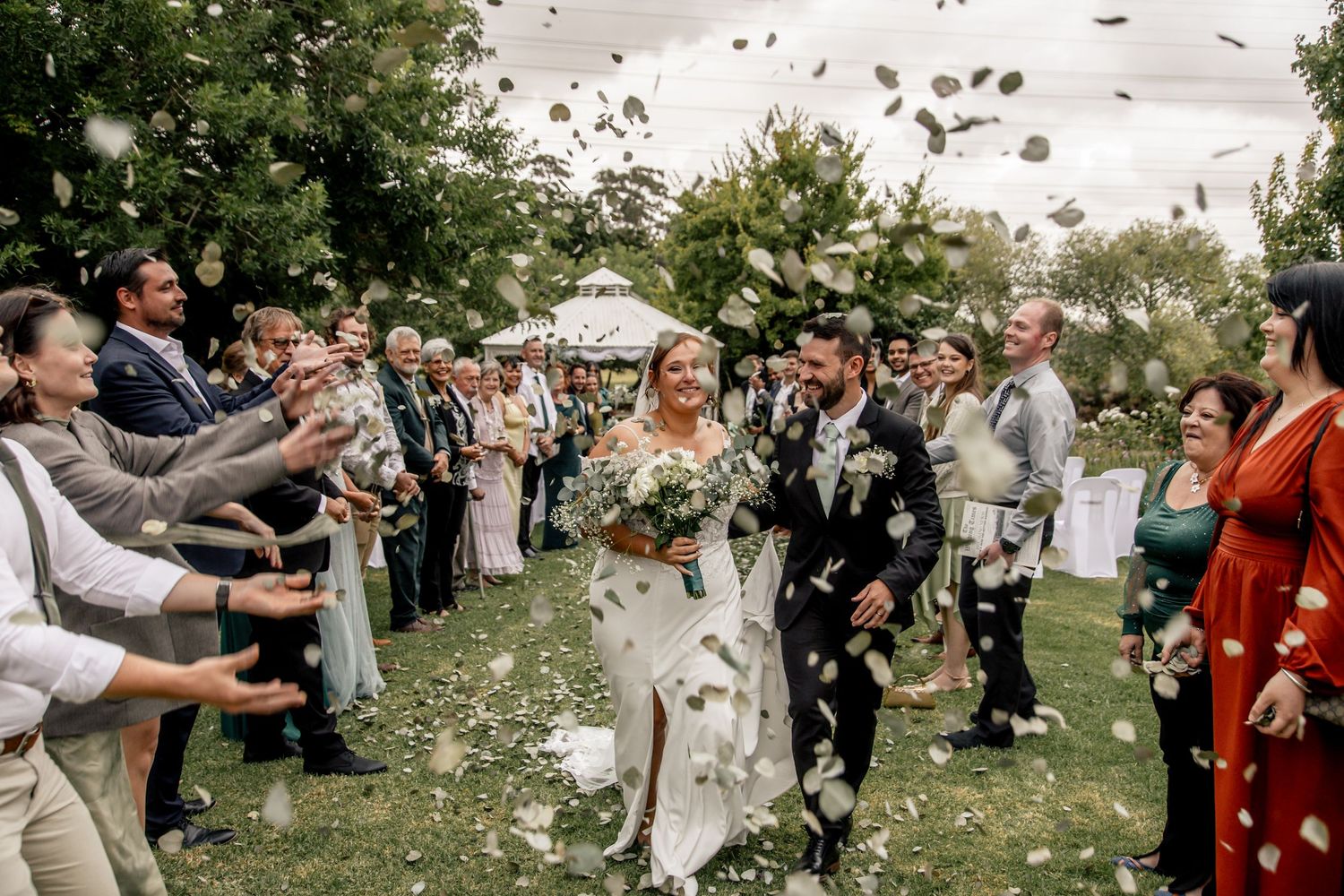 Bride and groom walk through confetti shower surrounded by cheering wedding guests outdoors.
