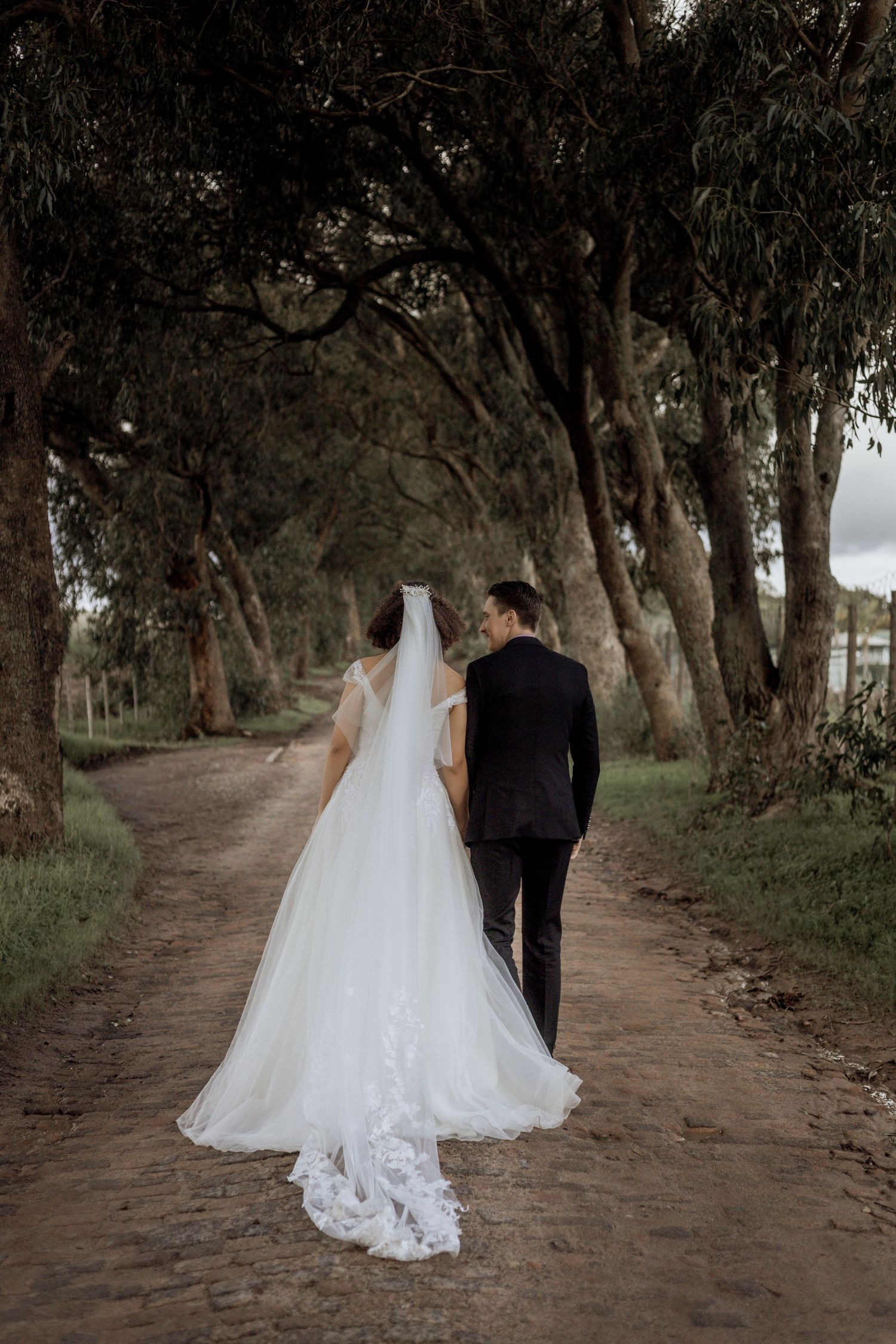Couple in wedding attire walking down a scenic tree-lined dirt path in a romantic outdoor setting.