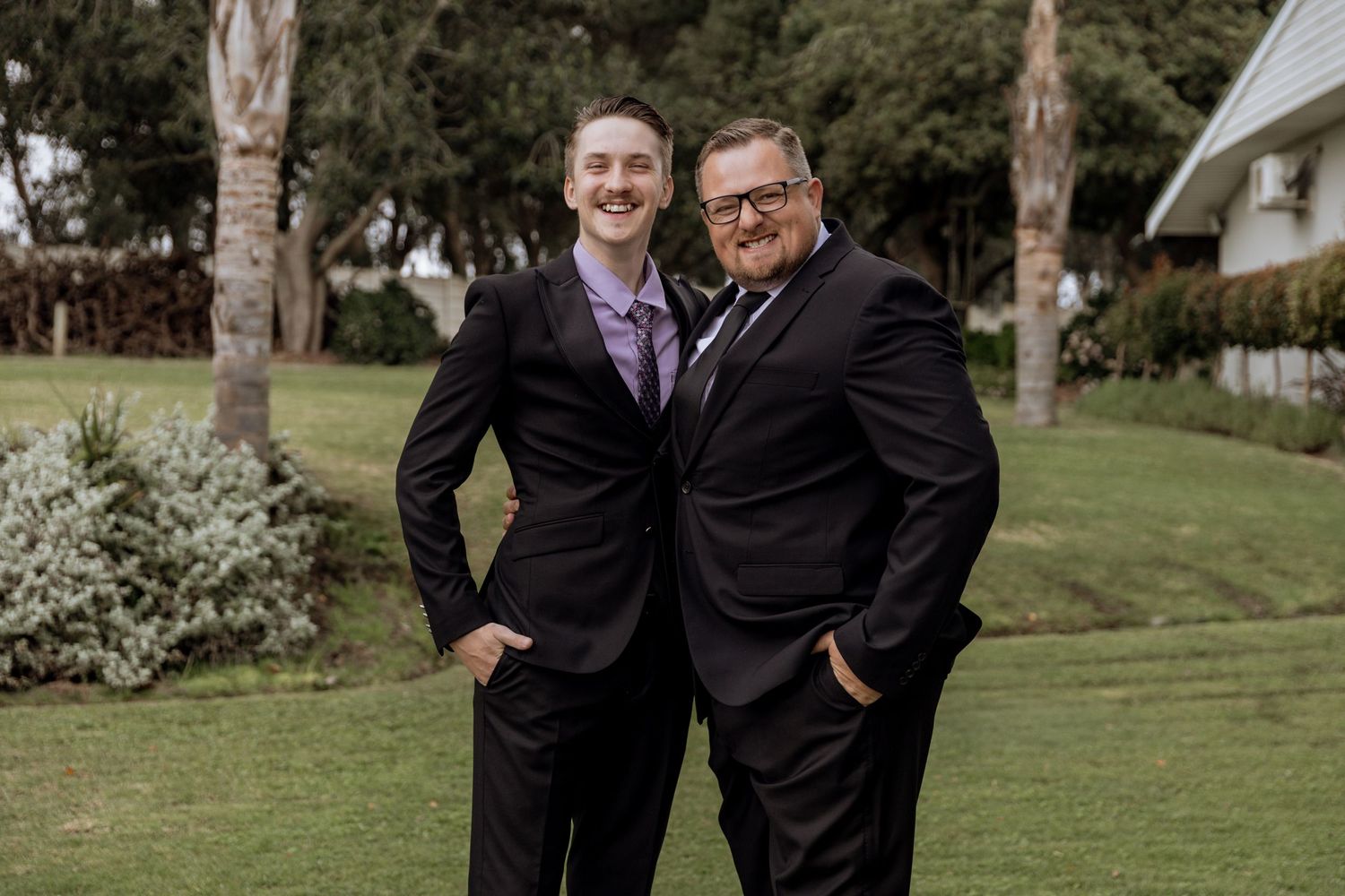 Two people in dark suits stand together in a garden setting while sharing a happy moment outdoors.
