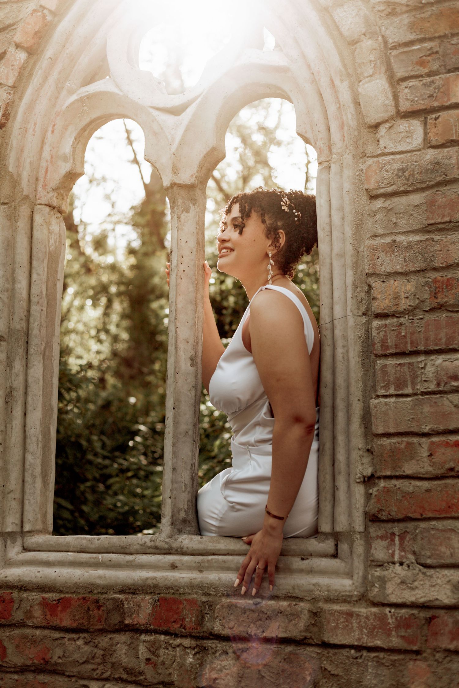A figure stands in a sunlit gothic stone archway wearing a white dress, with soft backlit glow filtering through the window.