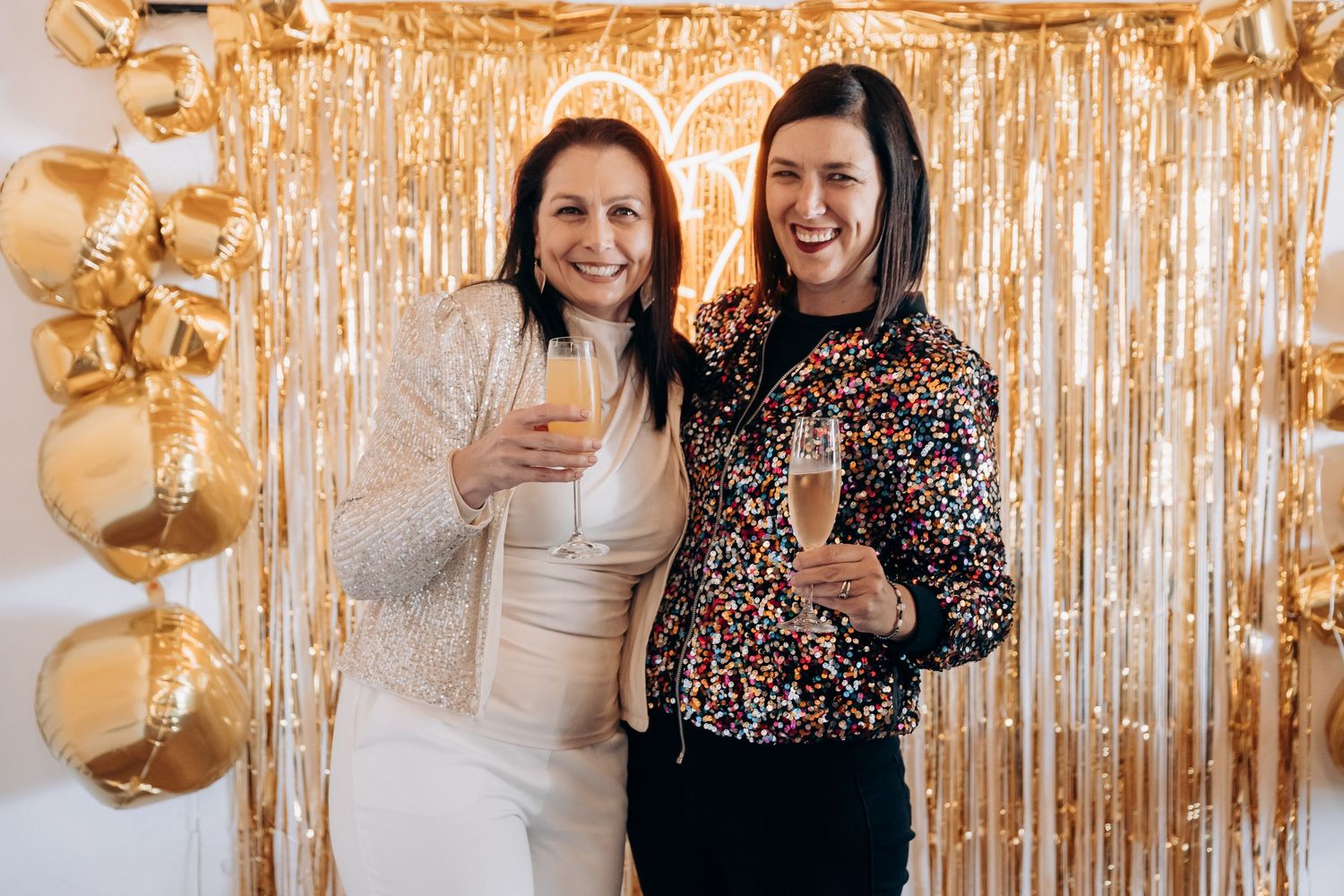 Two women smiling and holding champagne glasses in front of a gold tinsel backdrop with balloon decorations.