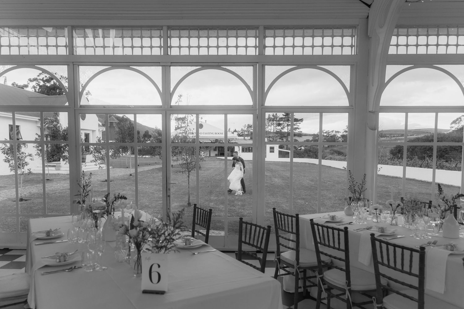 Black and white photo of an elegant wedding reception hall with arched windows, set tables, and guests visible outside.