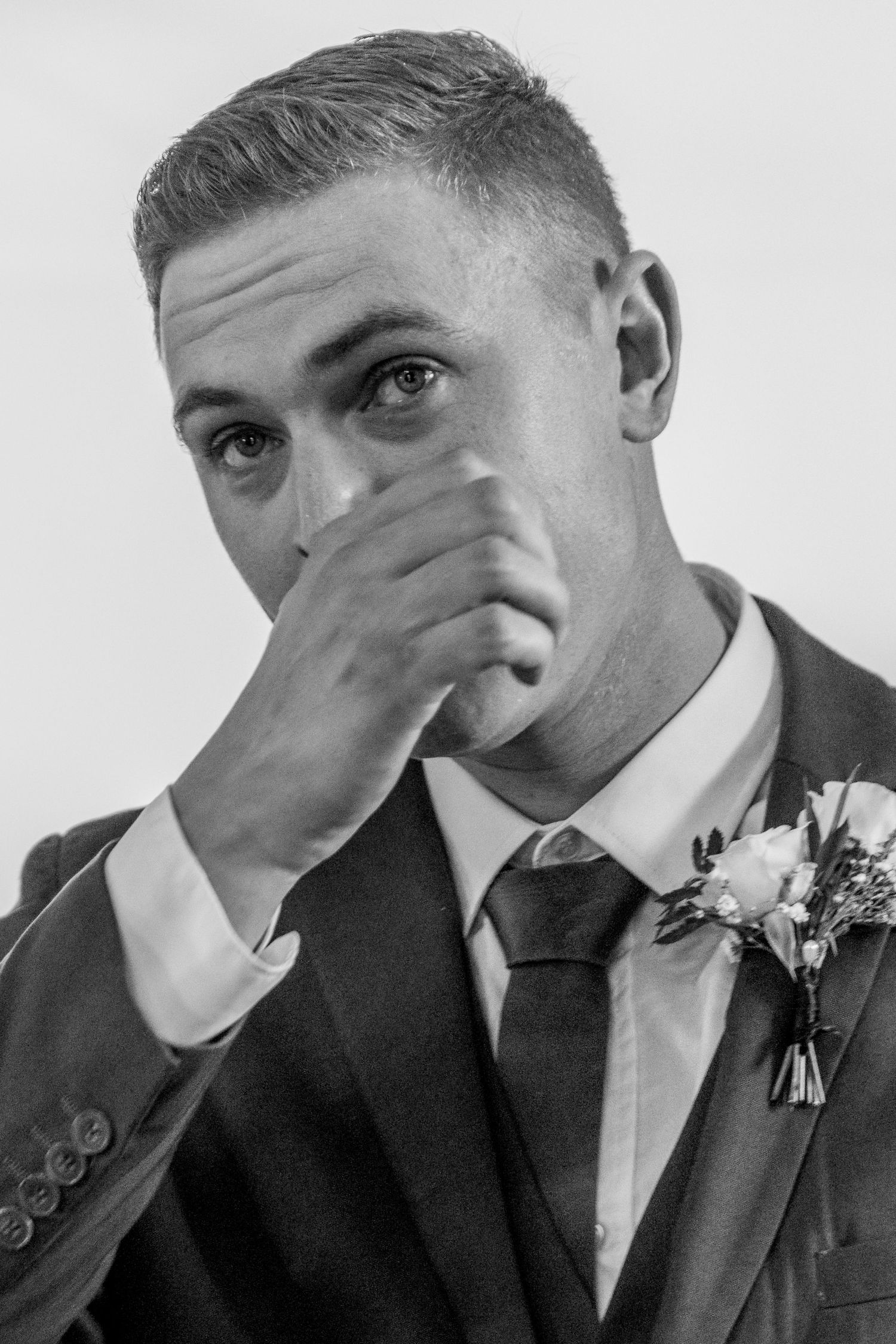 Black and white photo of emotional groom in suit with boutonniere covering mouth with hand at wedding.